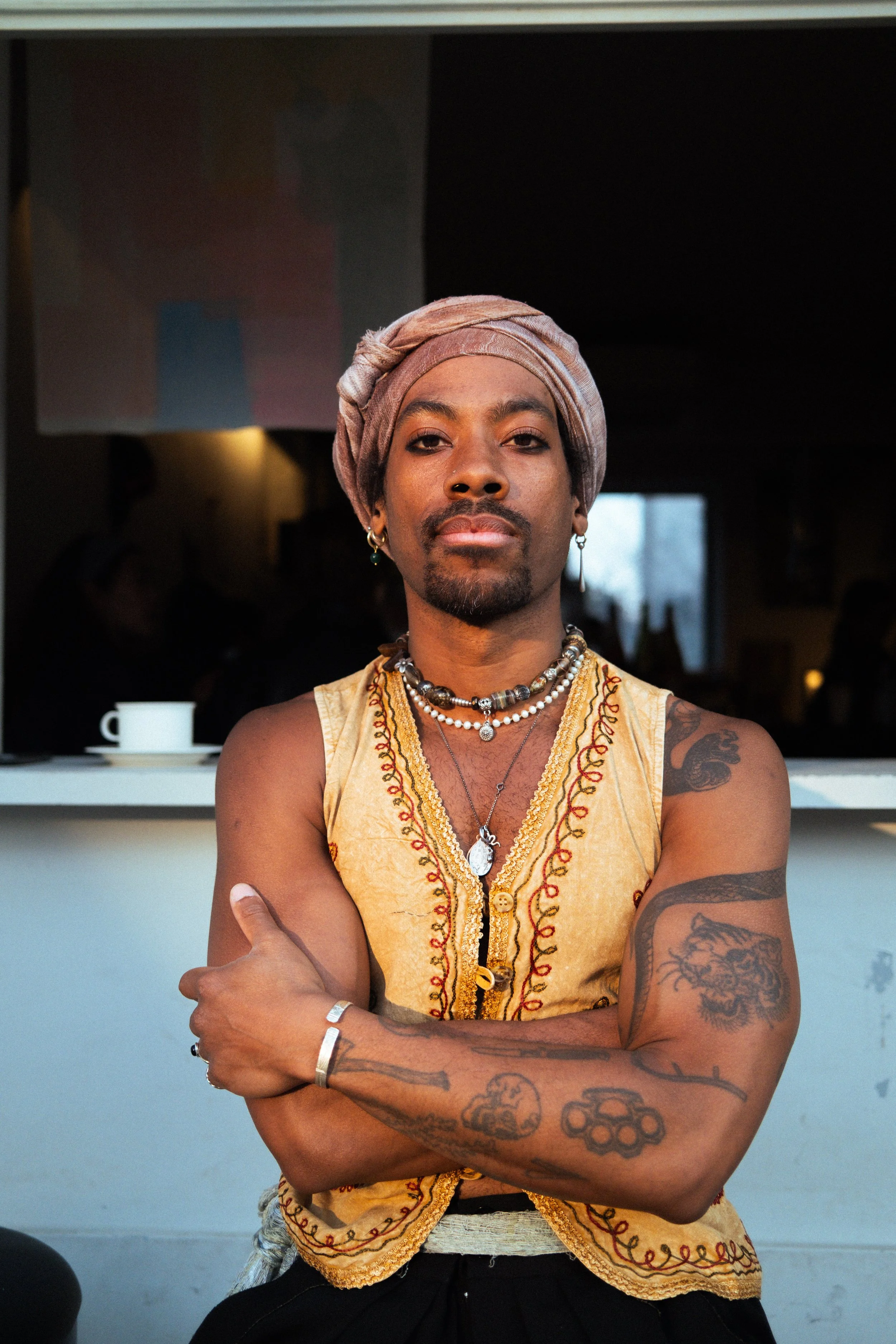 A person with dark skin and tattoos on both arms, wearing a beige sleeveless top with embroidery, layered necklaces, earrings, and a headwrap, sitting with arms crossed in front of a background featuring a window and a coffee cup.