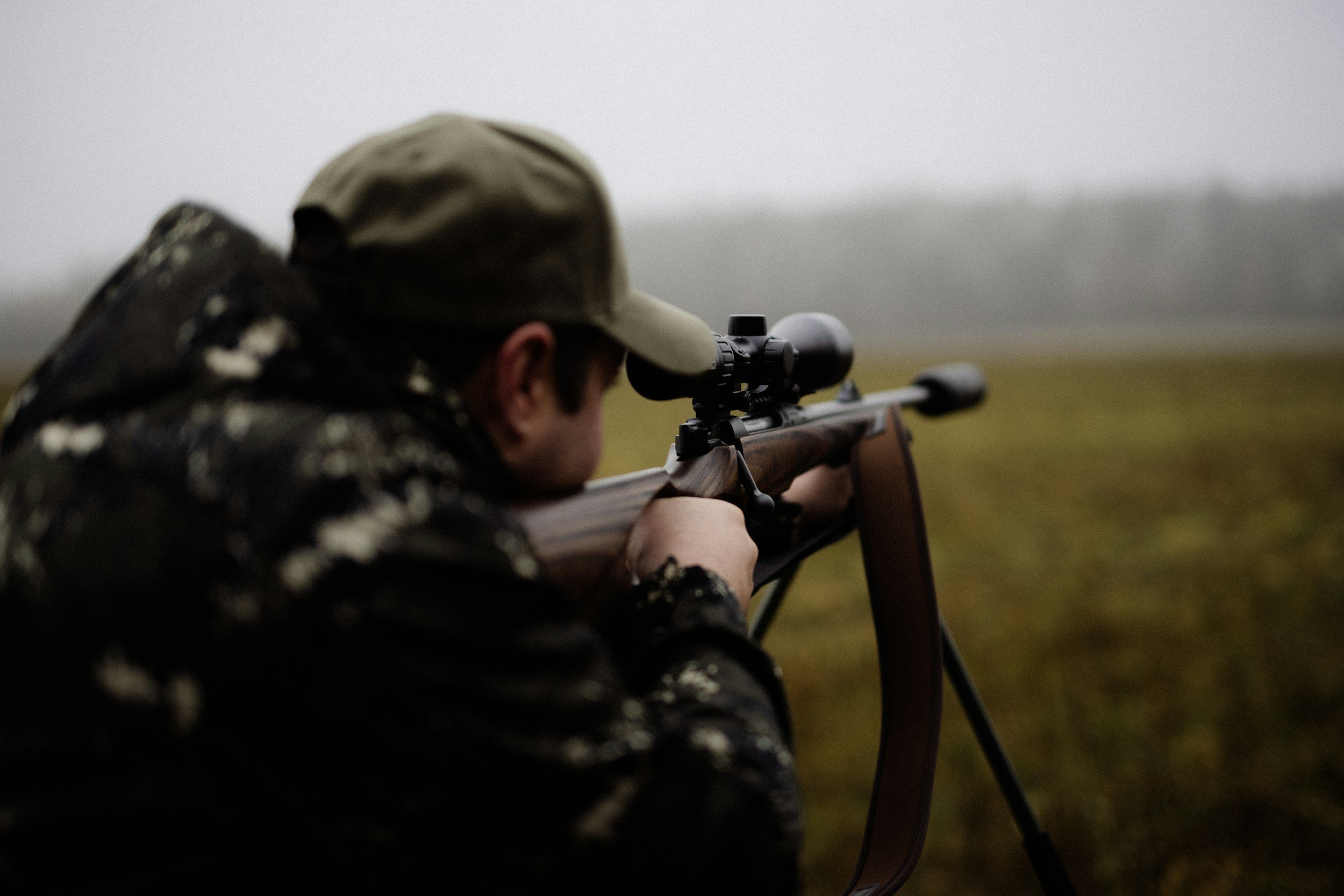 A person wearing camouflage clothing and a cap, lying down and aiming a scoped rifle outdoors in a foggy or overcast environment.