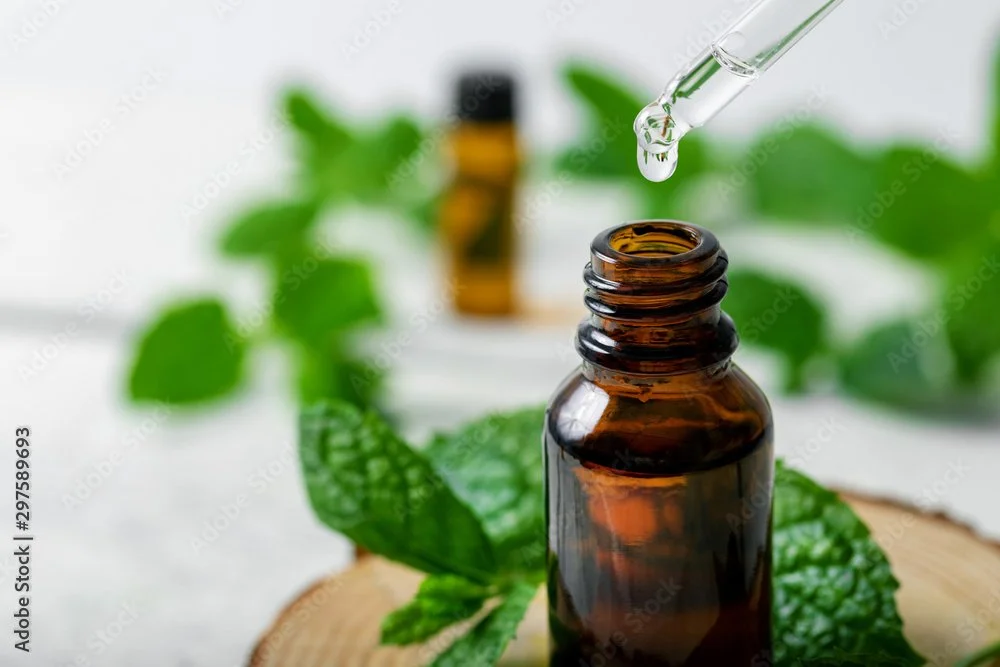 A brown glass bottle with a dropper pipette releasing a liquid, surrounded by green leaves, set on a wooden surface, with another small bottle blurred in the background.