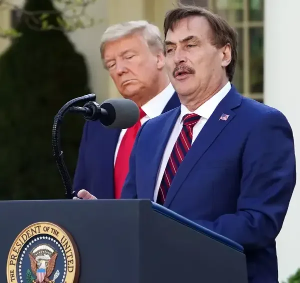 A man with a mustache speaking at a podium outdoors, with former President Donald Trump standing behind him, both in suits and ties.