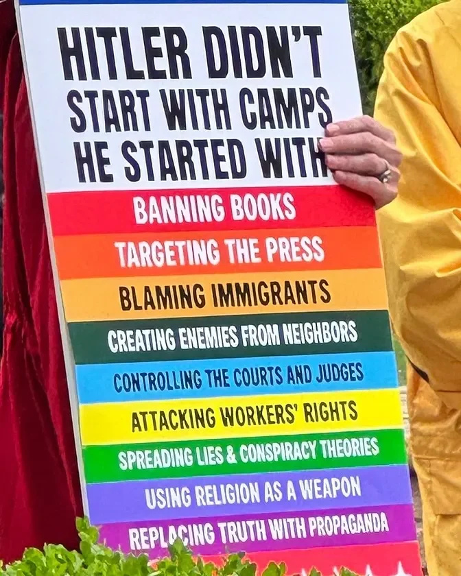 A protest sign with the patriotic colors of the American flag, listing claims about what Hitler started with, including banning books, targeting the press, blaming immigrants, creating enemies from neighbors, controlling courts and judges, attacking workers' rights, spreading lies and conspiracy theories, using religion as a weapon, and replacing truth with propaganda.