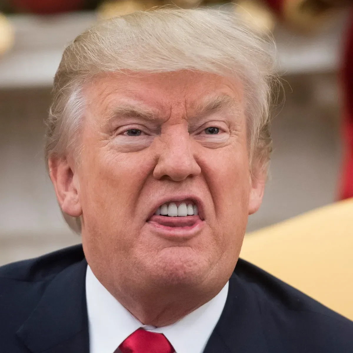Close-up of Donald Trump making a grimace with his mouth partly open, showing teeth, wearing a suit and red tie.