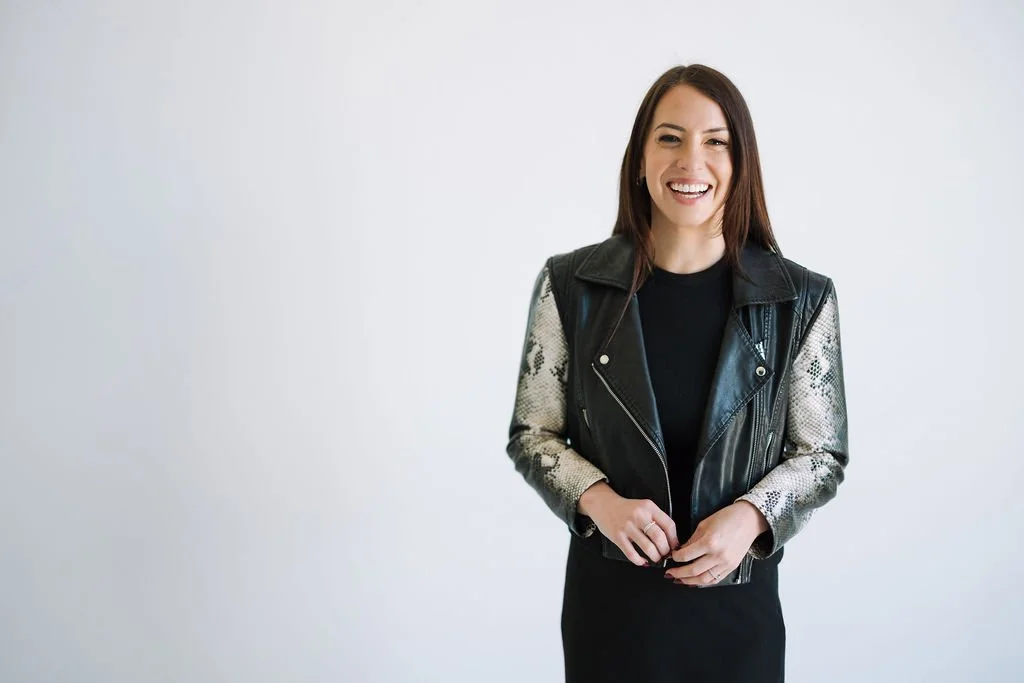 A smiling brunette, Elise Kennedy, with long hair and a grey background looks at the camera