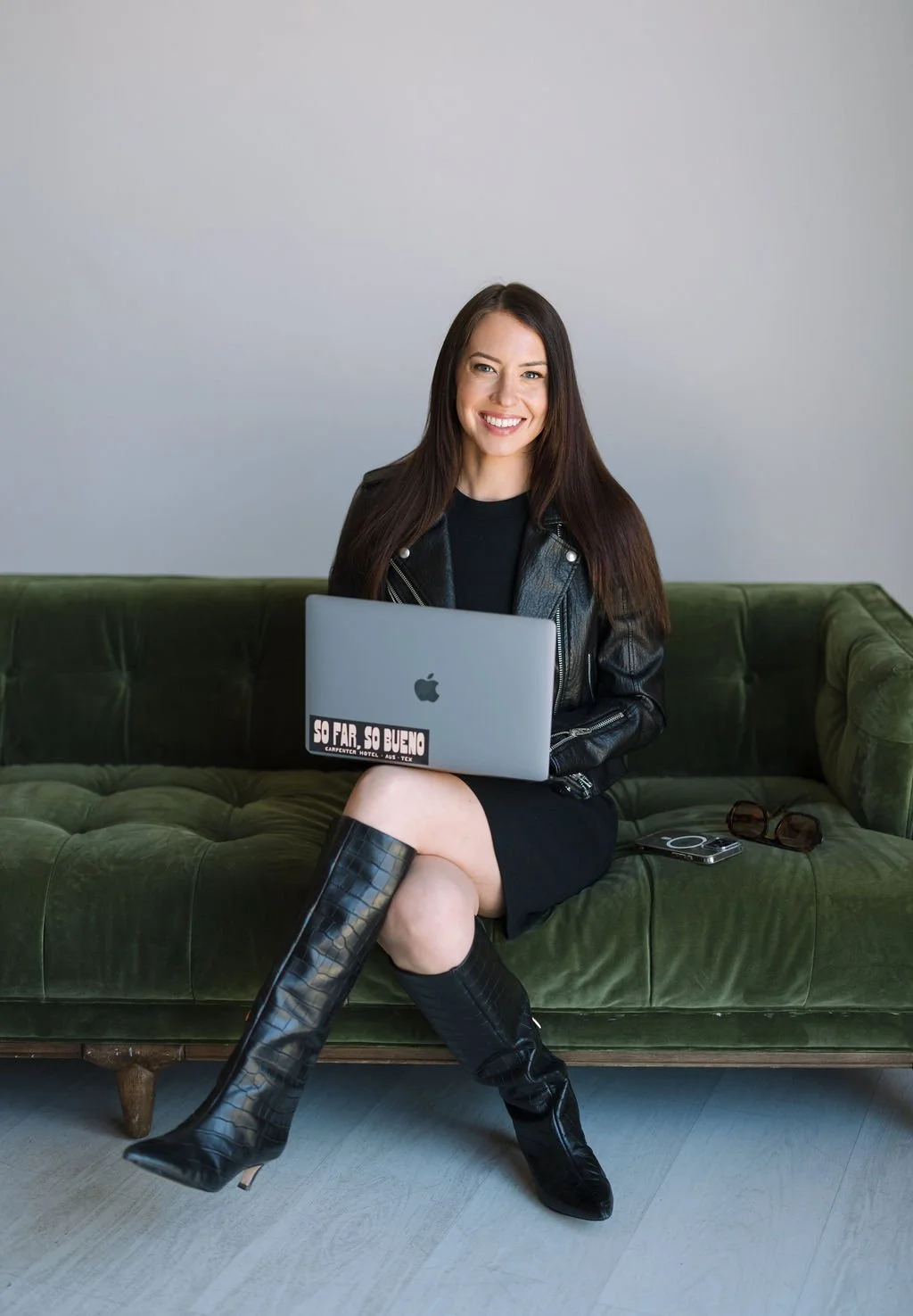Smiling woman with long brown hair sitting on a green velvet couch, holding a silver laptop with a sticker that says "SO FAR, SO BUENO." She is wearing a black dress, a black leather jacket, and knee-high black leather boots. Sunglasses and a smartphone are on the couch beside her.