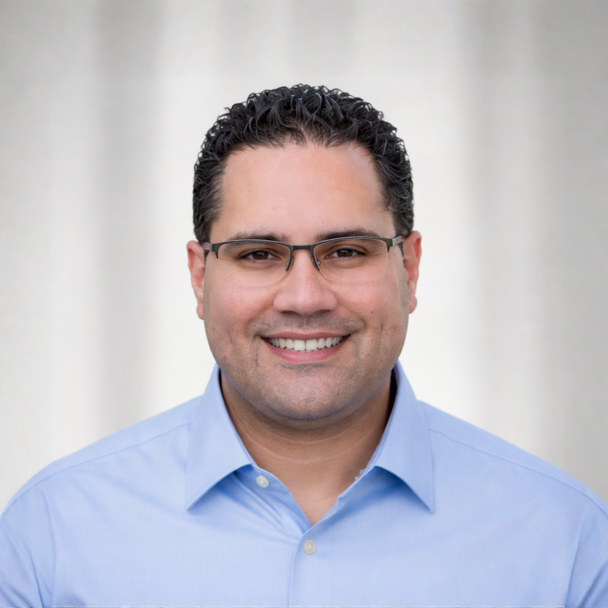 Close-up of a smiling man with black curly hair, wearing glasses and a light blue collared shirt.