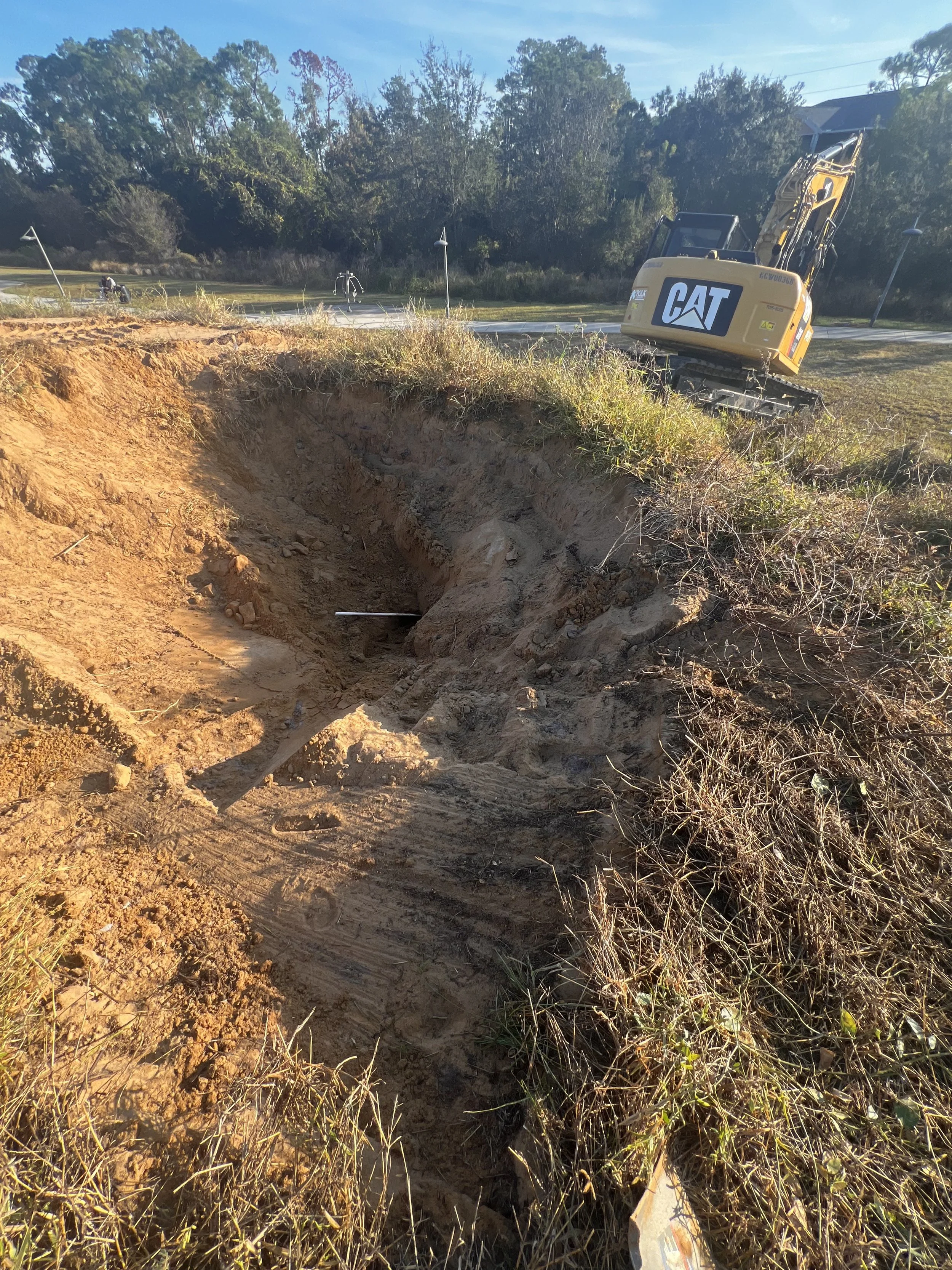 Excavation site with a pile of dirt and a small yellow CAT excavator on a grassy area with trees in the background.