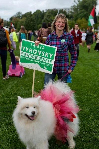 Tanya holding a Tanya Vyhovsky for State Senate sign behind her dog Laika who is wearing a hot pink tutu