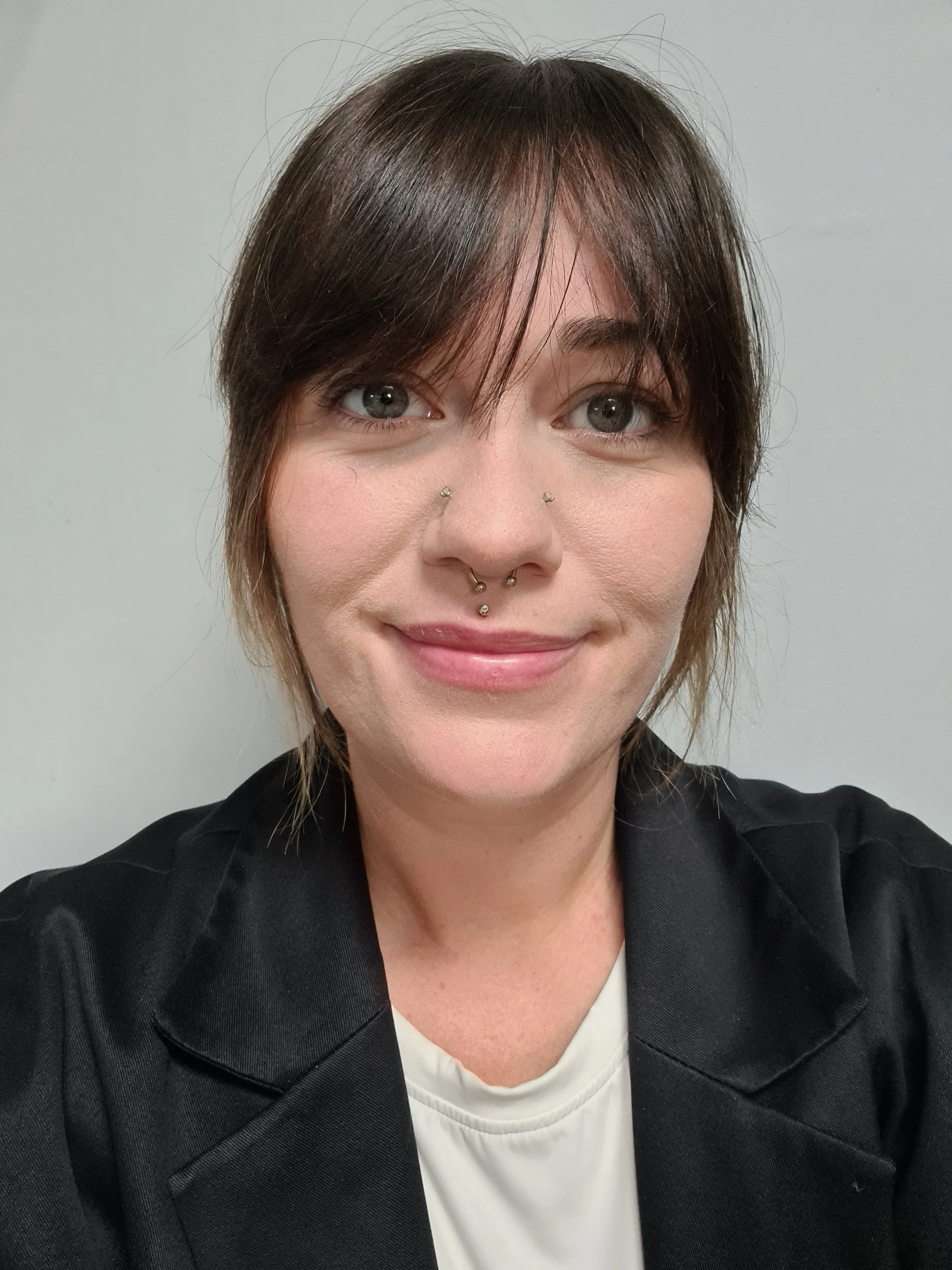 A woman taking a selfie in front of a plain, light-colored wall. She has short, layered brown hair, gray eyes, light skin, and is wearing a black jacket or coat over a white top. She has facial piercings including a septum ring and two nostril studs.
