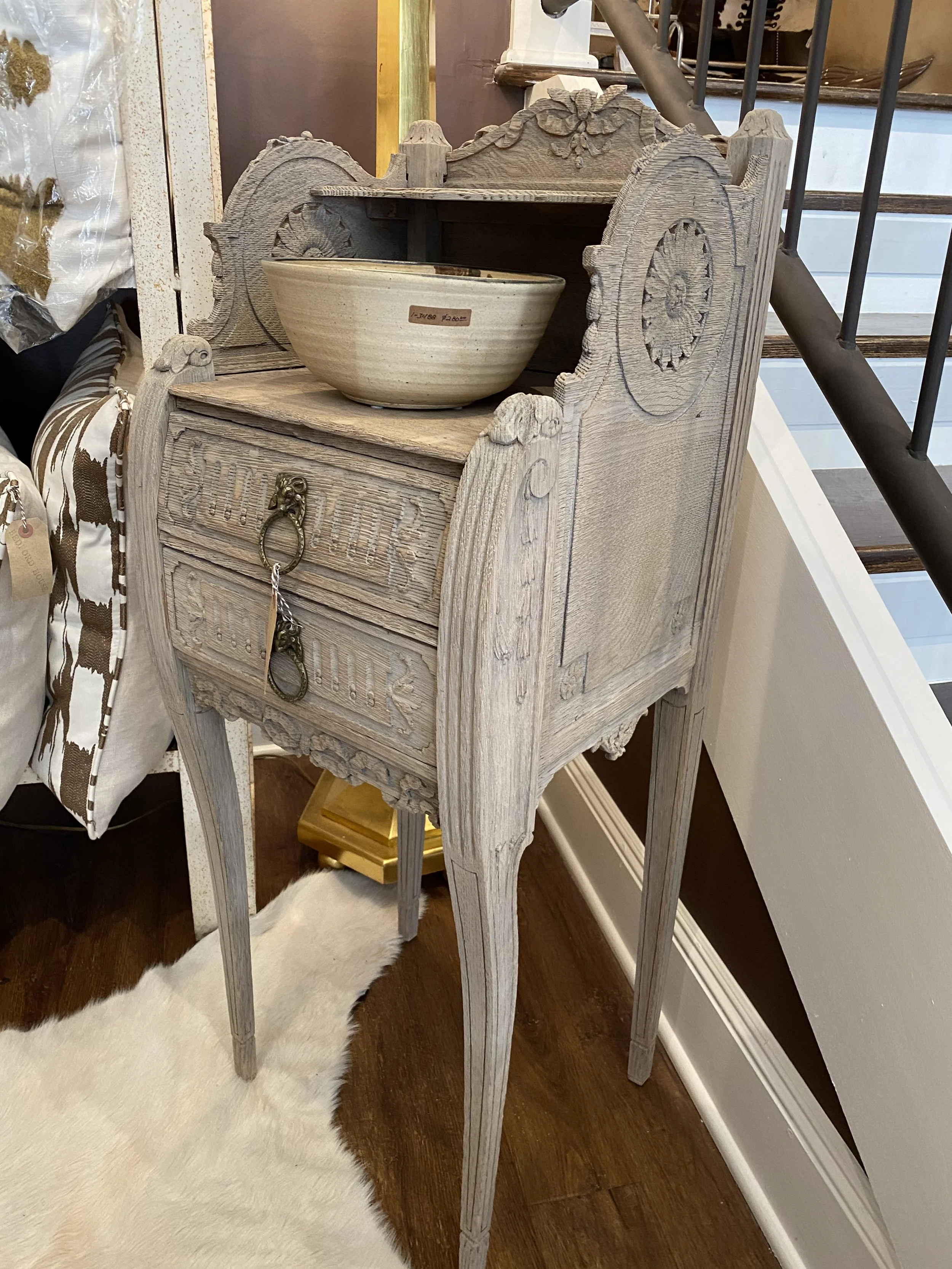 A small wooden cabinet with carved decorative details and a ring pull handle, topped with a ceramic bowl, standing on a dark wooden floor next to a white plush rug.