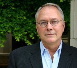 A middle-aged man with glasses and gray hair, wearing a dark suit and white shirt, standing outdoors with green trees in the background.