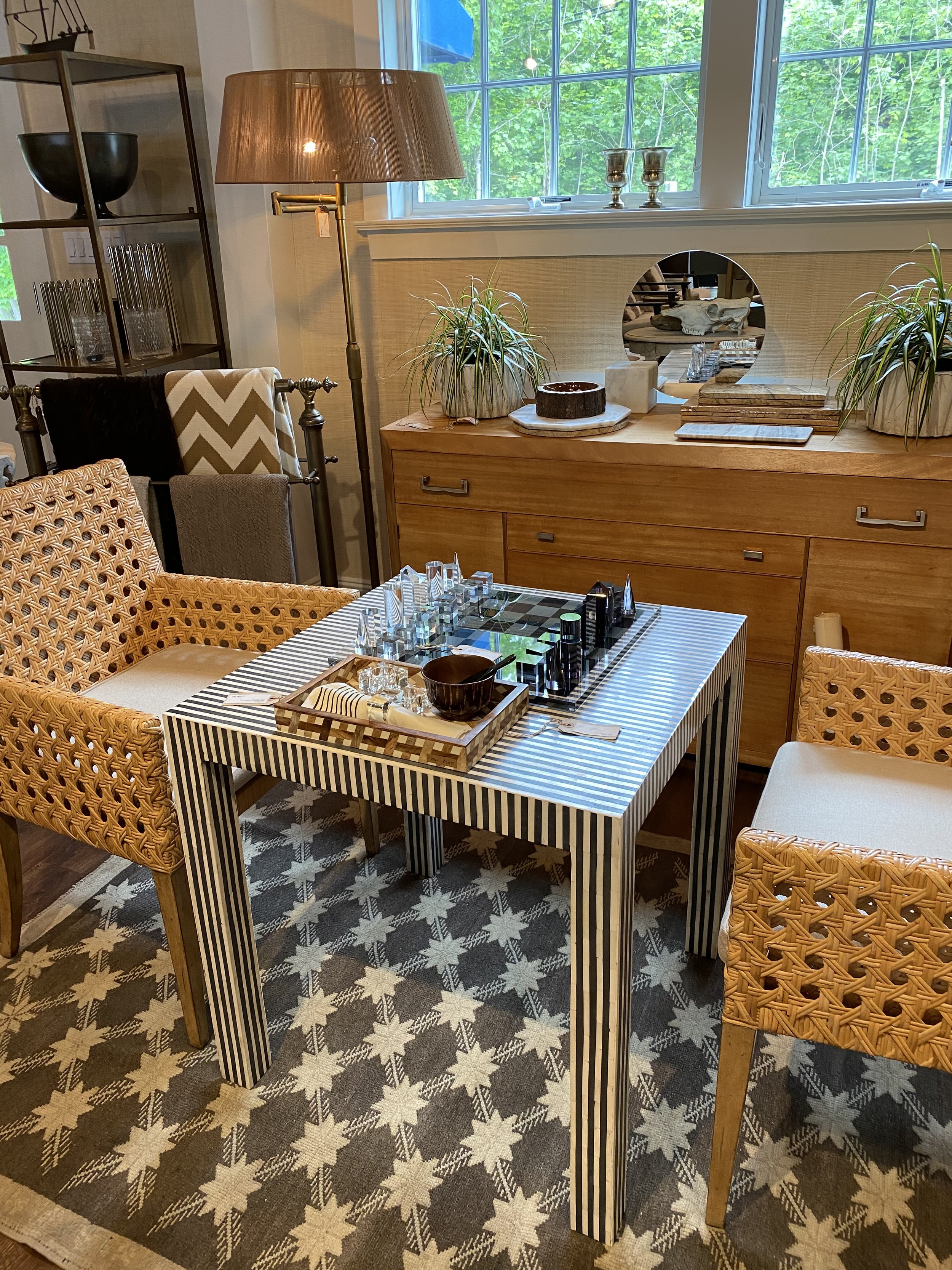 A dining room with a table that has a black and white striped pattern. The table is set with glasses, a dark bowl, and a tray with utensils. Surrounding the table are woven chairs and upholstered armchairs. Behind the table, a wooden sideboard holds 
