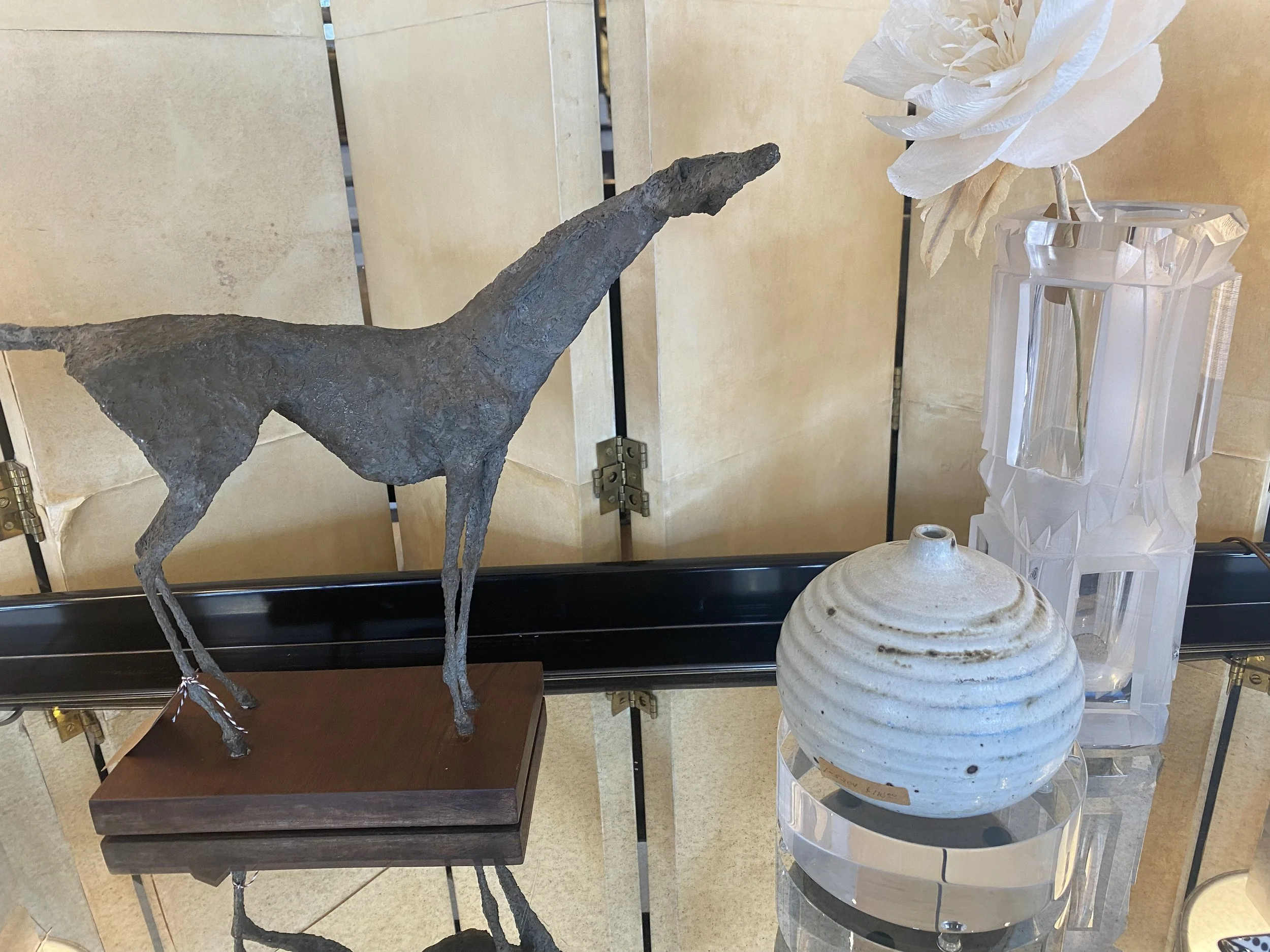A display of decorative items including a gray, textured horse sculpture on a wooden base, a white pleated paper flower in a faceted glass vase, and a white ceramic urn, all arranged on a tiered black and glass stand.