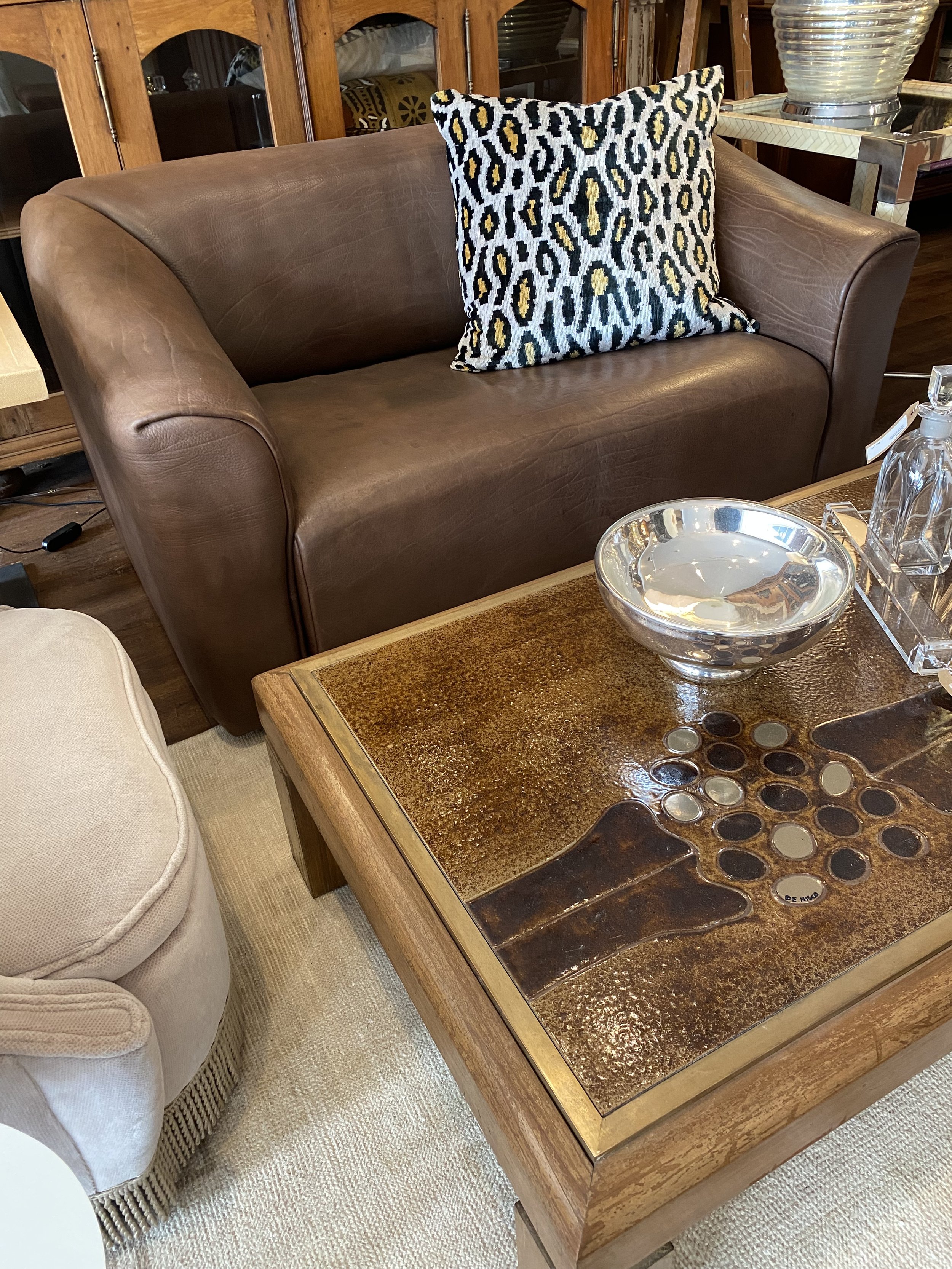 A living room scene showing a brown leather couch and a wooden coffee table with decorative inlay. The table has a large silver bowl, glass bottle, and a glass tray. Behind the couch is a wooden cabinet and an upholstered armchair.