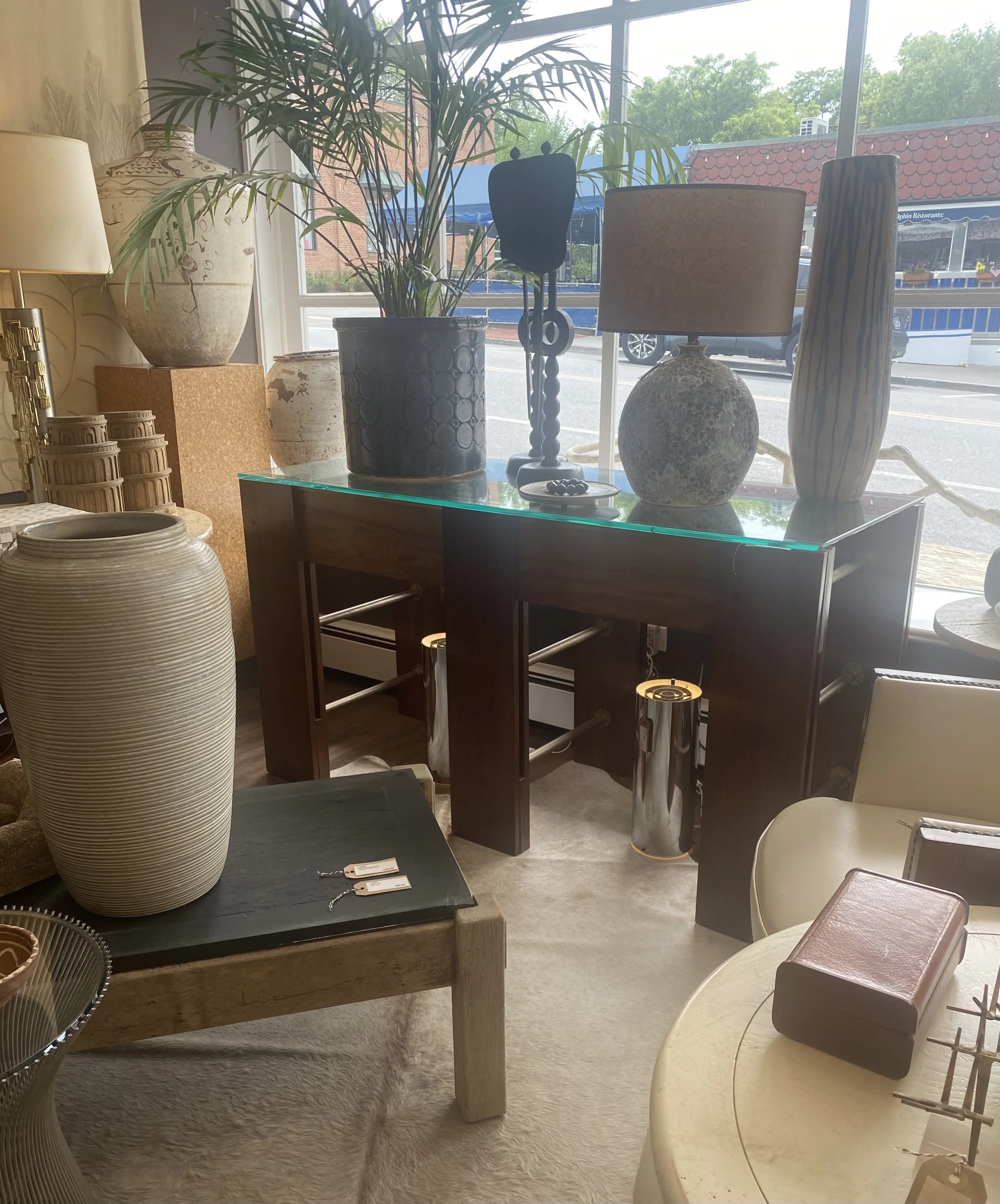 Interior of a furniture store with various decorative lamps and vases, a glass table, and other decorative items near large windows showing a street view outside.