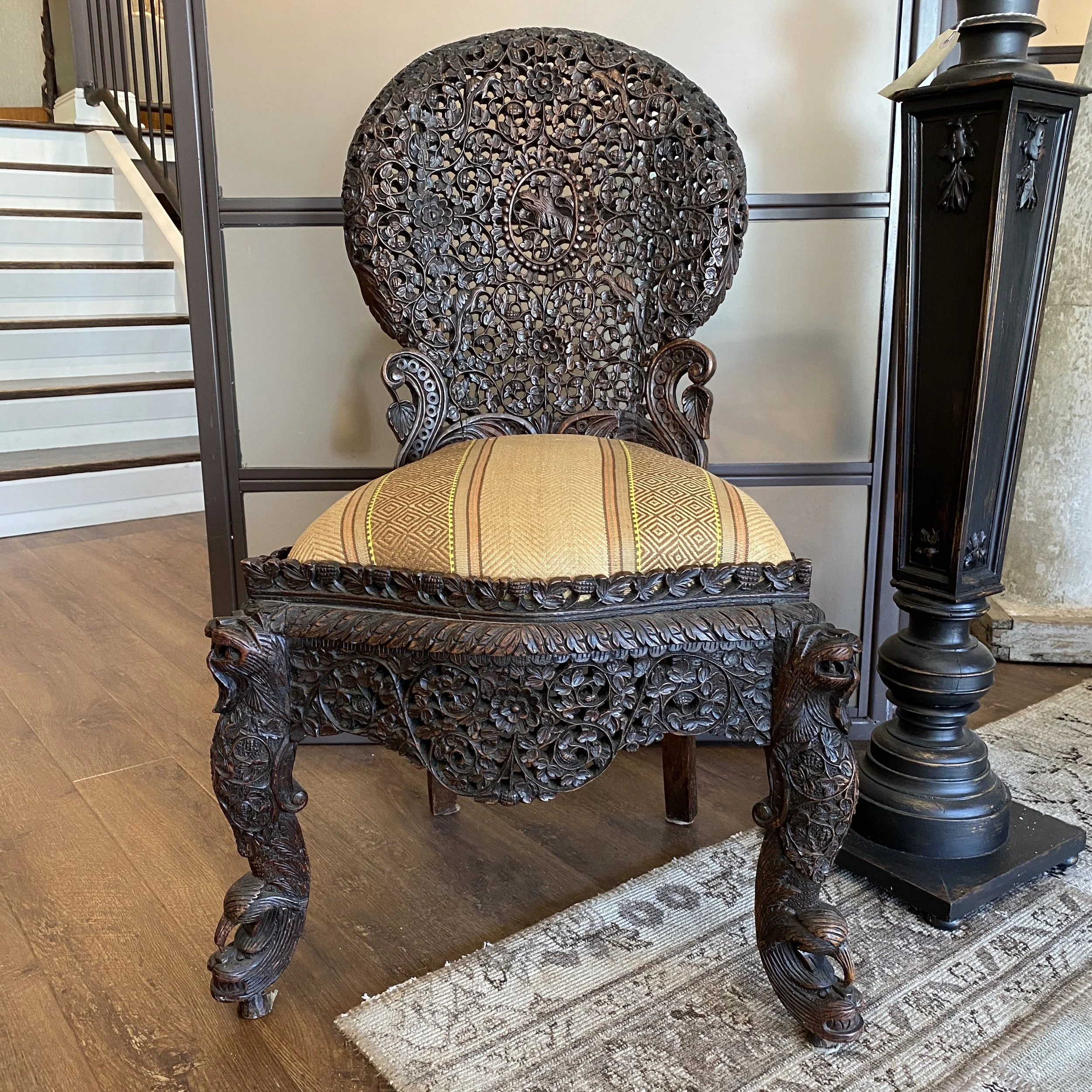 An intricately carved dark wooden chair with a tall, ornate backrest featuring floral and leafy patterns. The chair has a cushioned, yellow and gold striped seat. To the right, there is a black pedestal with decorative carvings, topped with a small g