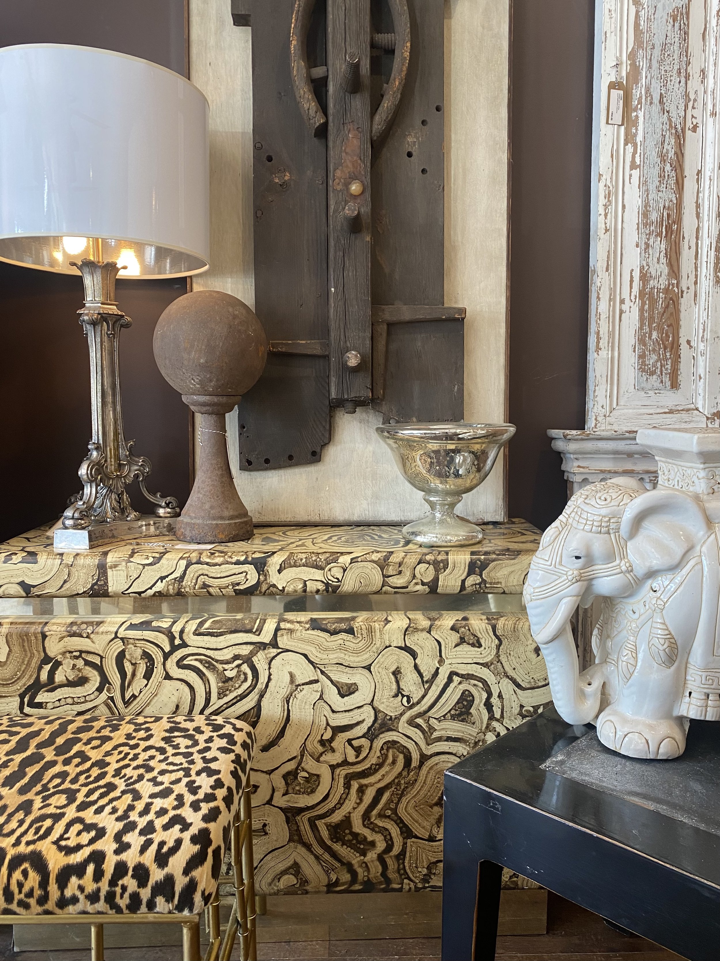 Decorative interior display including a leopard print stool, an ornate gold lamp, an abstract black and white patterned surface, a white carved elephant sculpture, and various antique or vintage furniture pieces including a wooden wheel and a distres