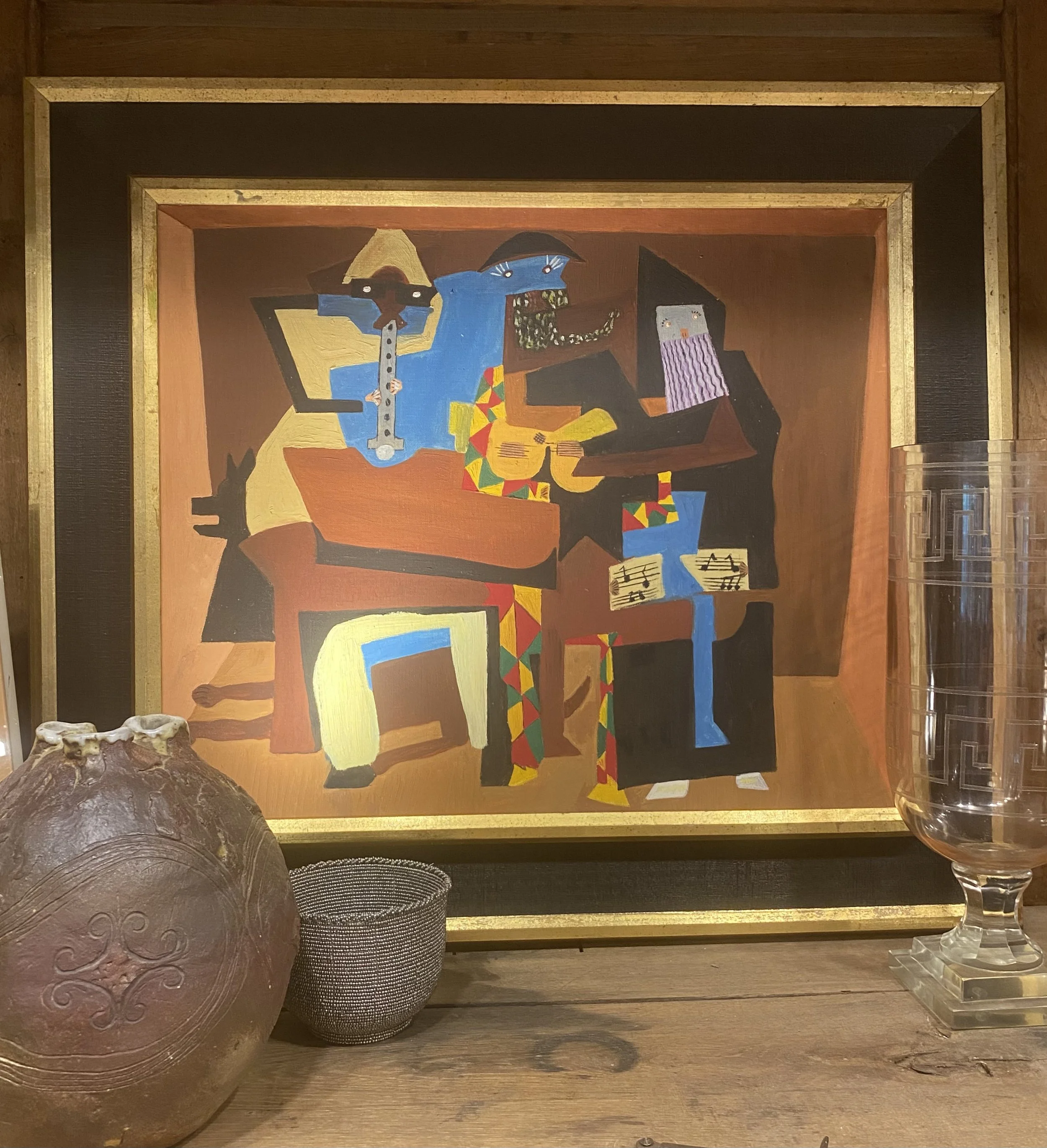 The image shows a colorful abstract painting of a person playing the guitar inside a room, as viewed from a table setting with decorative objects in the foreground, a metallic vessel and glass ashtrays, with a wooden surface and frame.