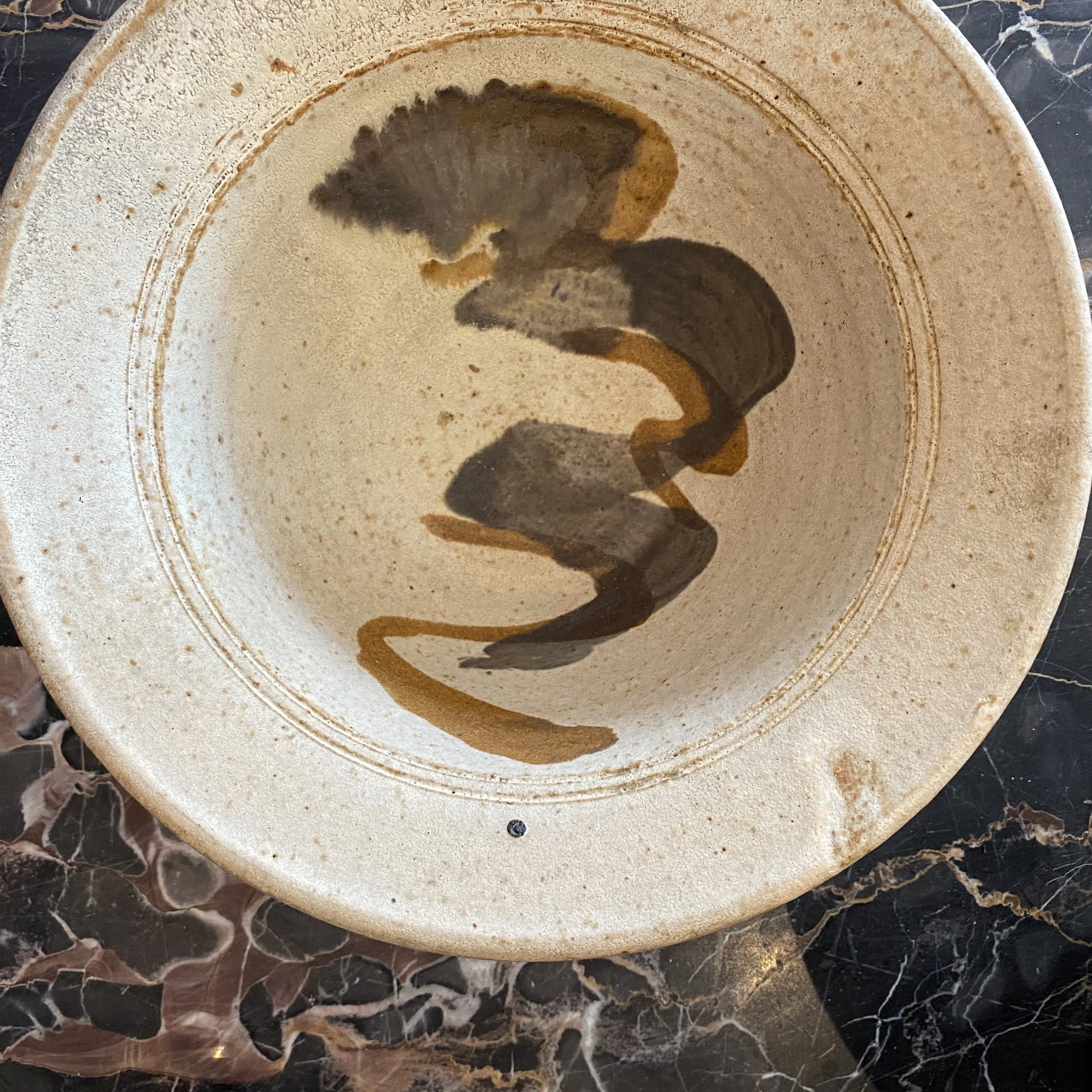 A top-down view of a round stone or ceramic plate with a brown design resembling a stylized dragon or mythical creature, placed on a black marble surface with white and pink veins.