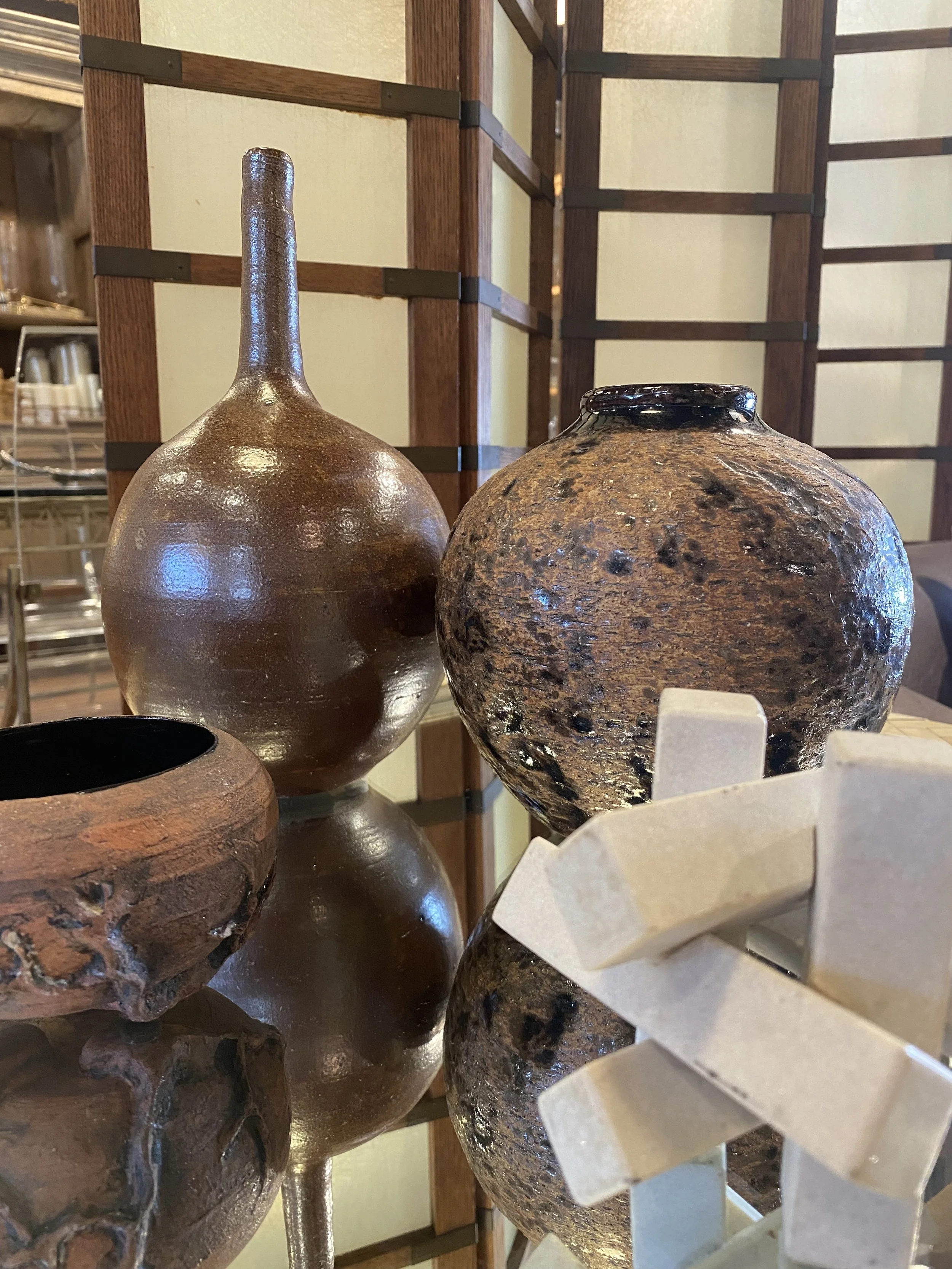 Two ceramic vases on display, one with a shiny, brown glaze and a narrow neck, and the other with a textured, earth-toned, rounded shape, seen in a store with wooden shelving and a decorative shelf in the background.