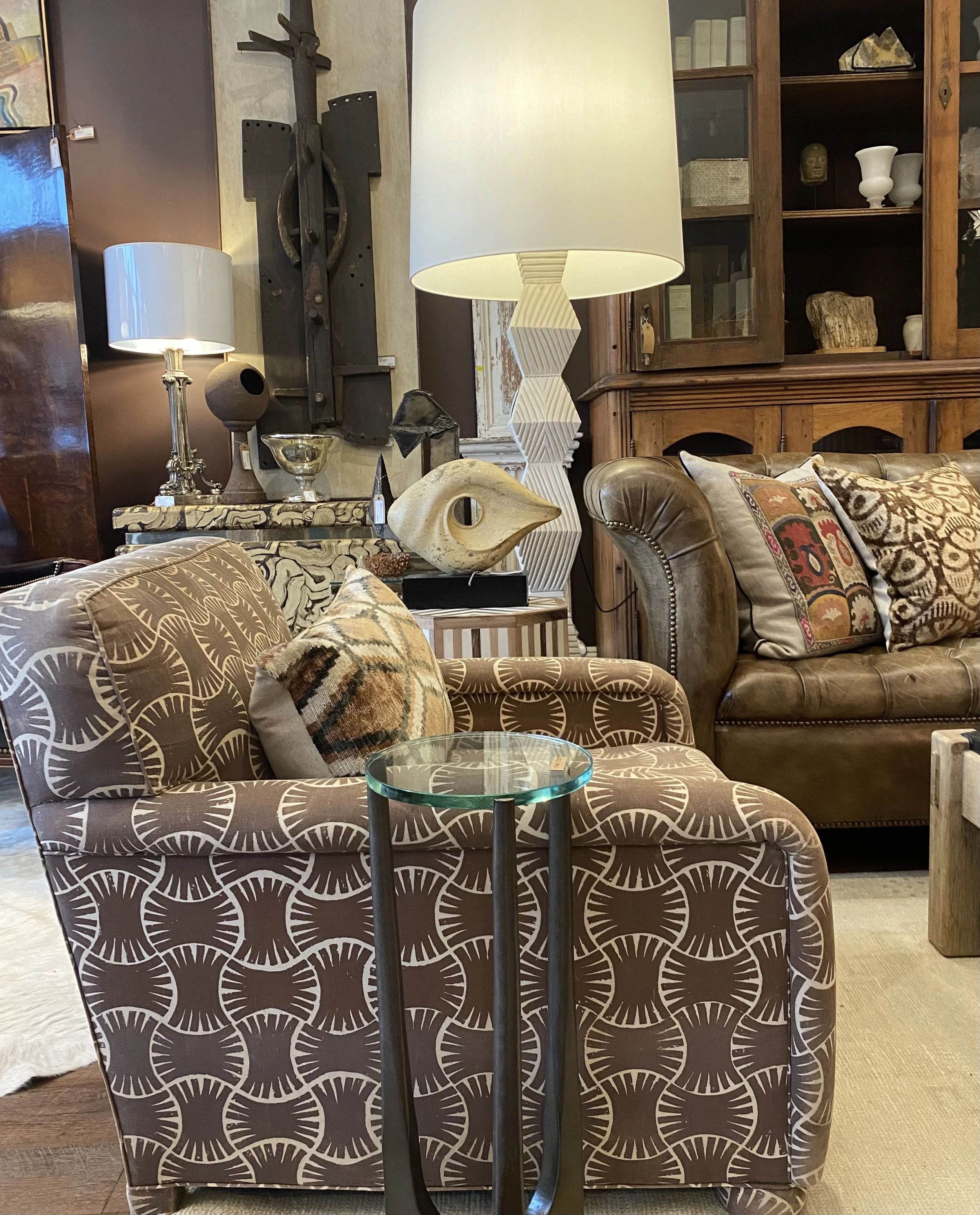 A living room with patterned sofa, decorative pillows, a glass-top side table, and various lamps. Background includes wooden shelves with decorative items and a wooden piece on the wall.