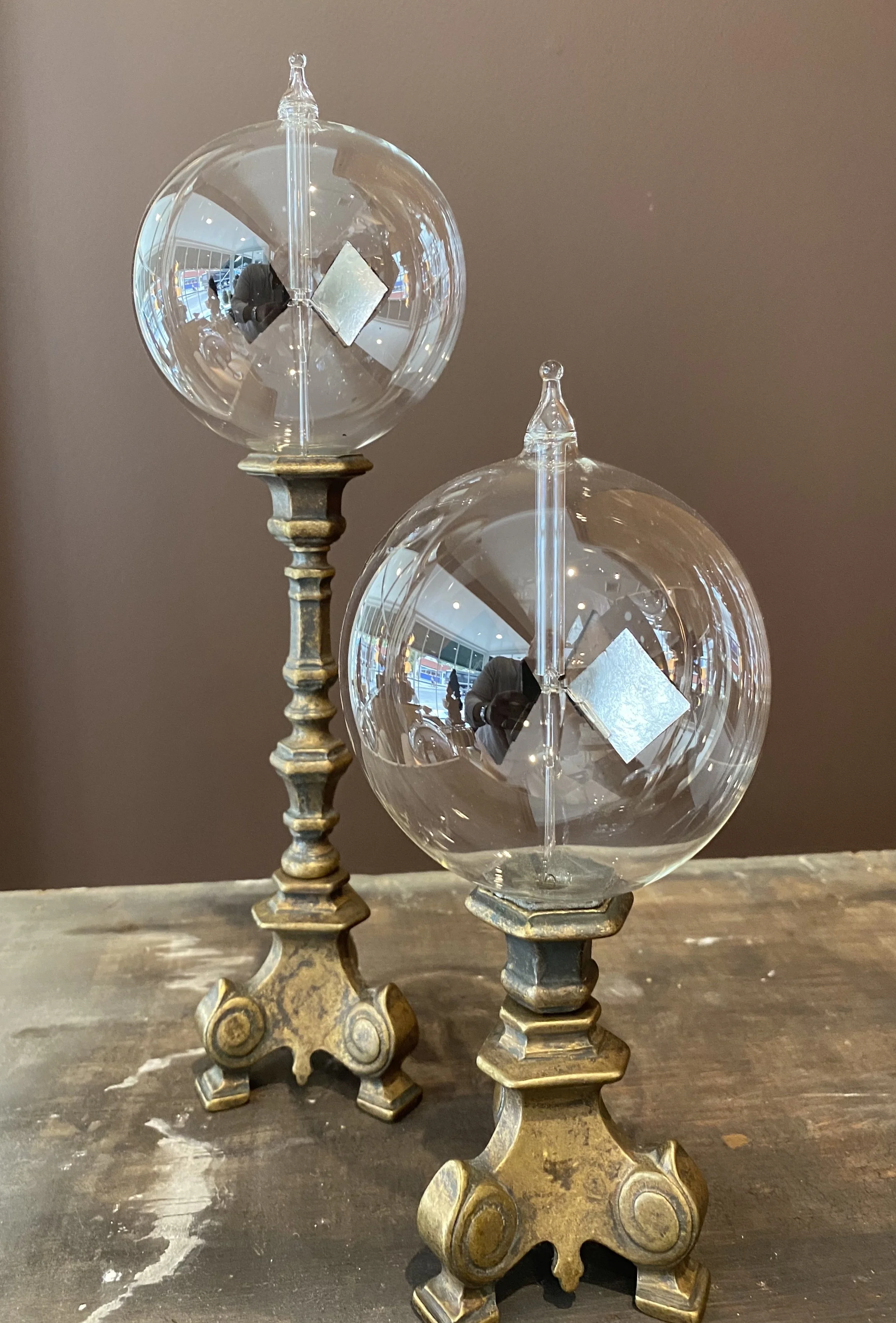 Two ornate brass candle holders with glass globes on top, placed on a wooden surface, with a brown background.