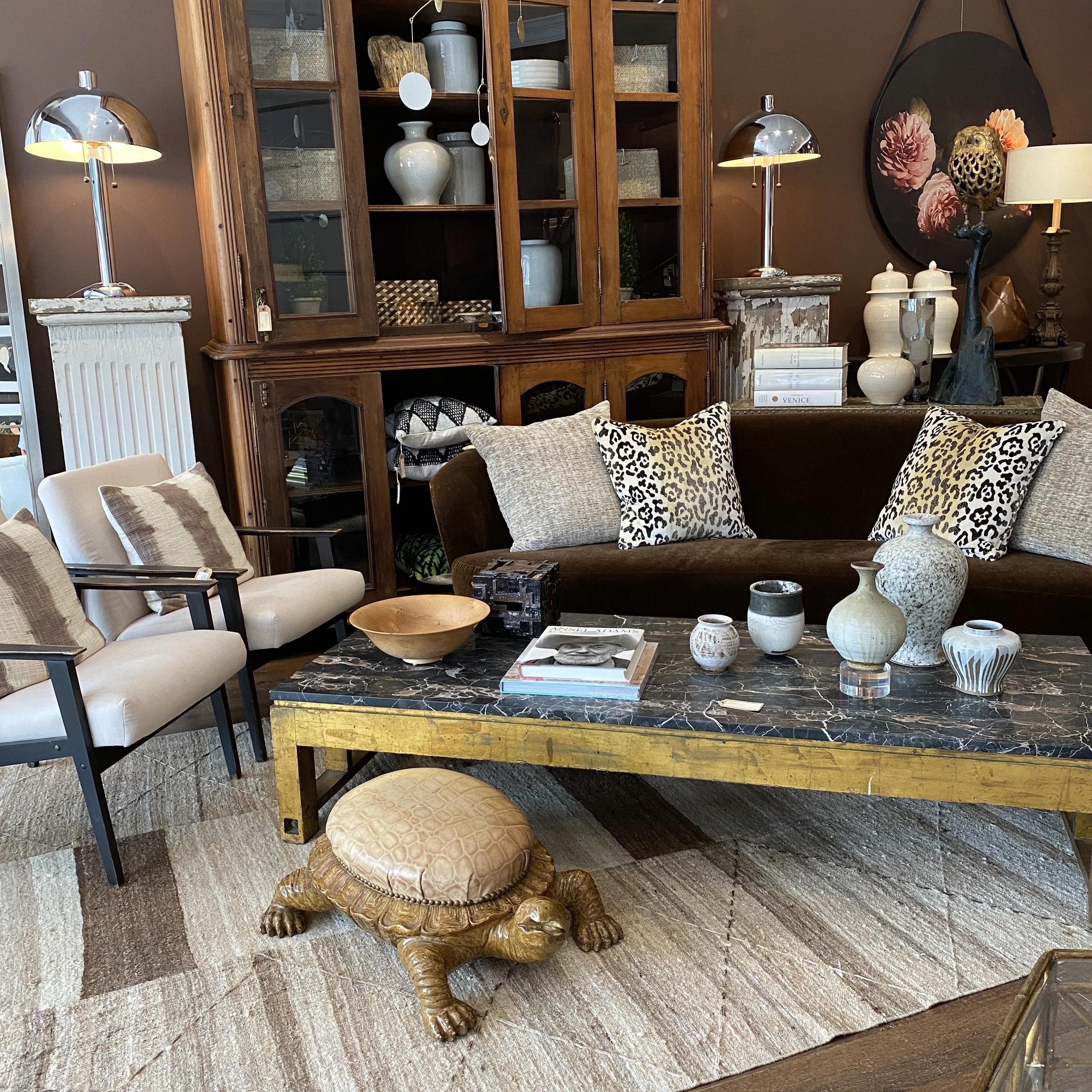 Living room with a wooden display cabinet filled with vases and baskets, standing against a brown wall. A black round floral wall art piece is on the wall, with a cream-colored table lamp and decorative items on a side table beneath it. To the right,