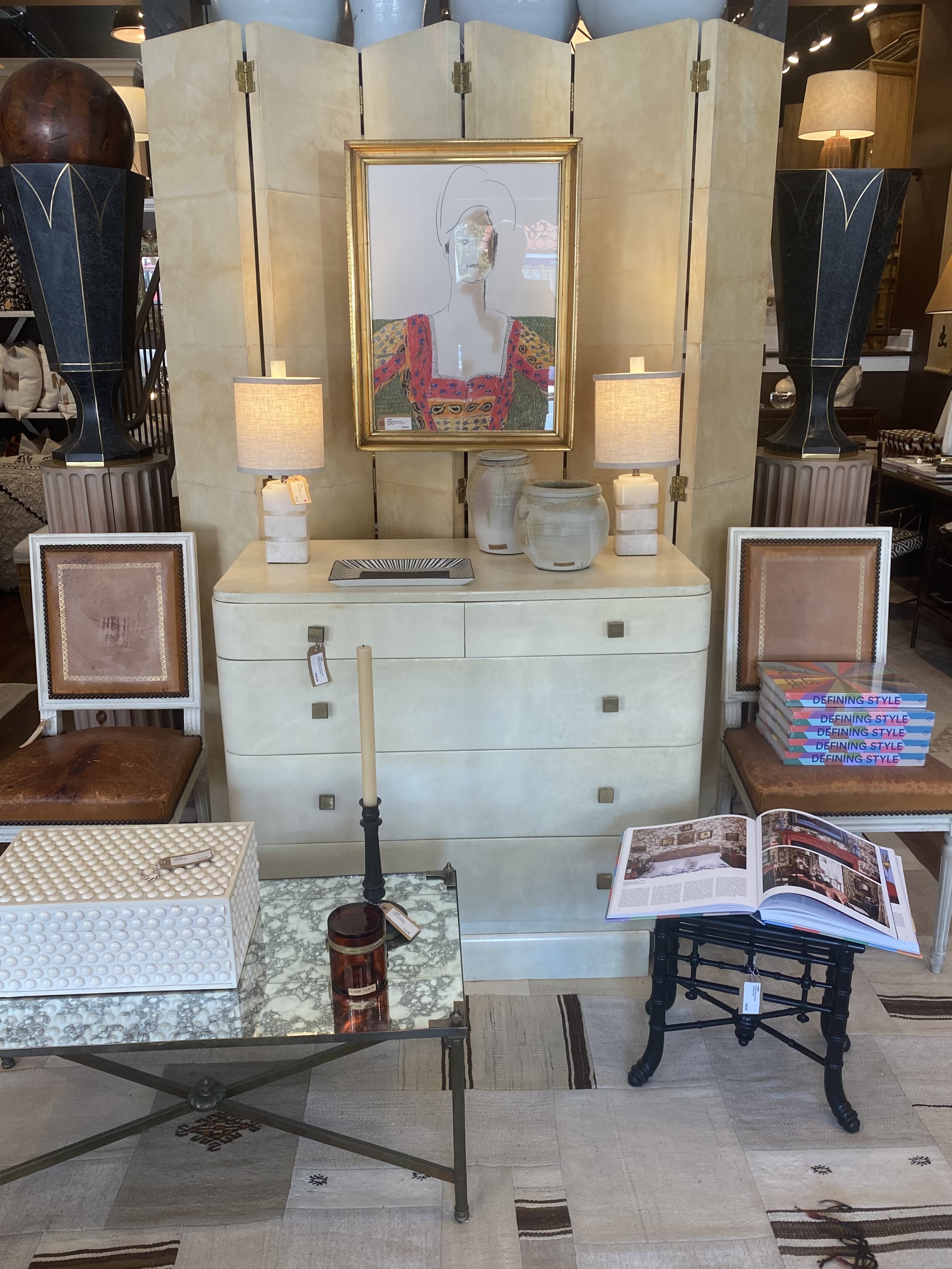 Various pieces of furniture and decor in a showroom including lamps, a framed sketch of a woman, vases, and tables with books. The items are arranged on and around a cream-colored dresser and display space.