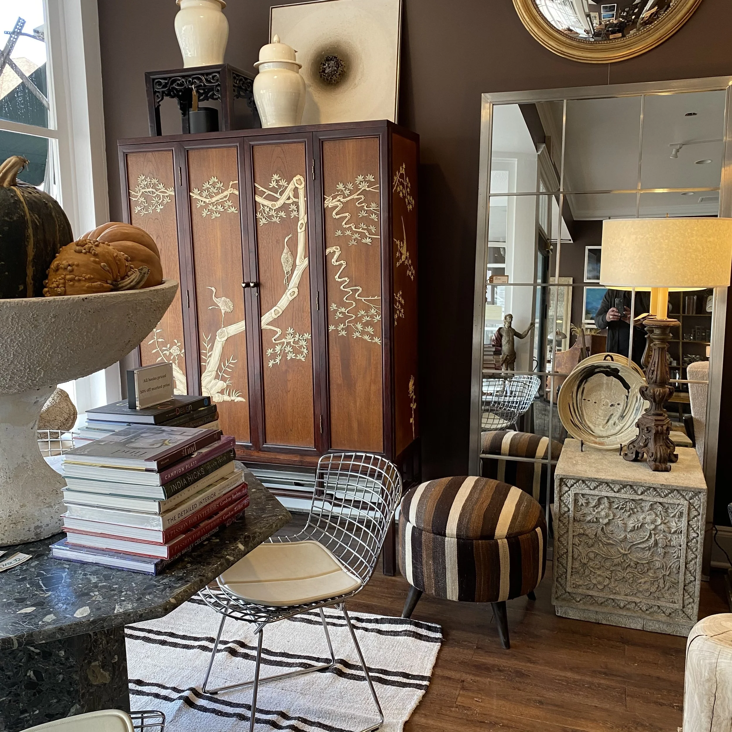 Interior of a furnishings or antique store with a wooden folding screen decorated with a painted crane and tree branches, surrounded by various decorative items including books, sculptural pieces, and ceramic objects. There is a large mirror reflecti