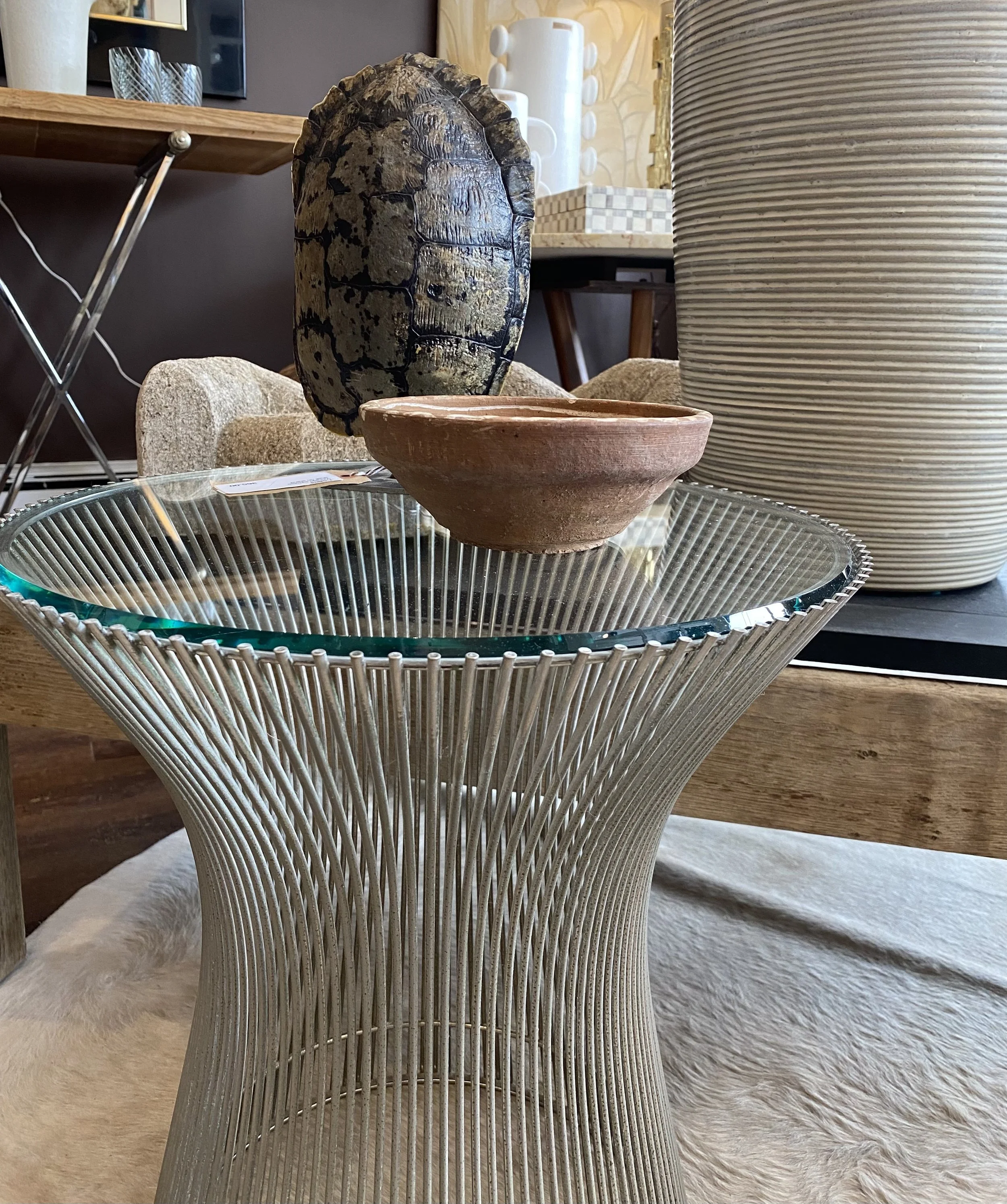 A room with various decorative objects including a wire bowl table, a large ceramic vase, a clay bowl, and a decorative sculpture of a turtle shell, with a wooden table and beige carpet in the background.