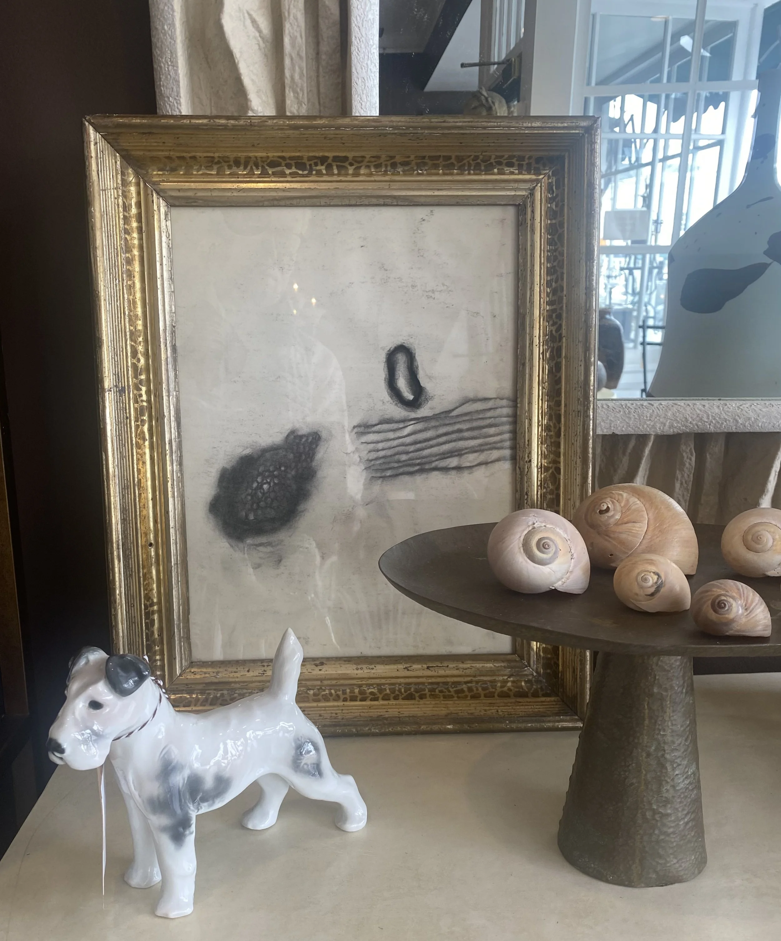 Display of a white ceramic dog sculpture, a group of seashells on a dark round table, a framed artwork or mirror with a gold patterned frame, and a grey cylindrical sculpture base in a store or gallery setting.