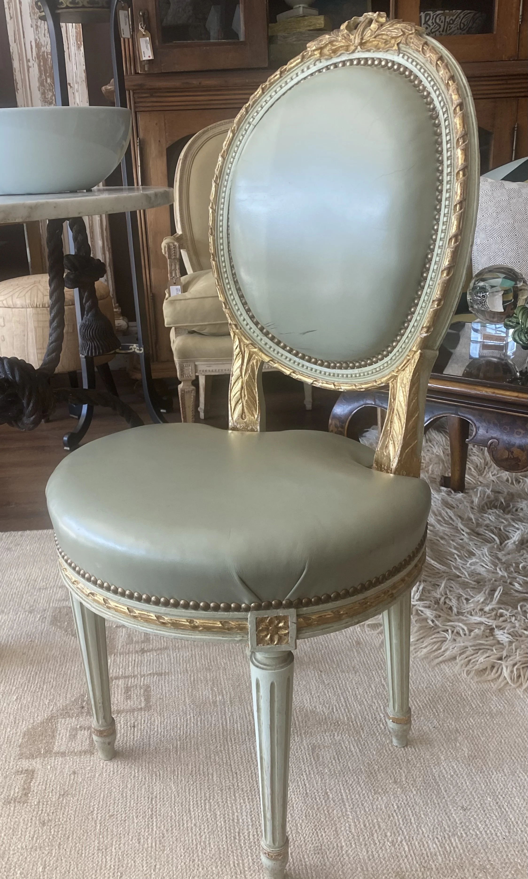 A vintage-style chair with a gold painted wooden frame and light green upholstered cushion and back. The chair is in a furniture store among other antique-style chairs and furniture.