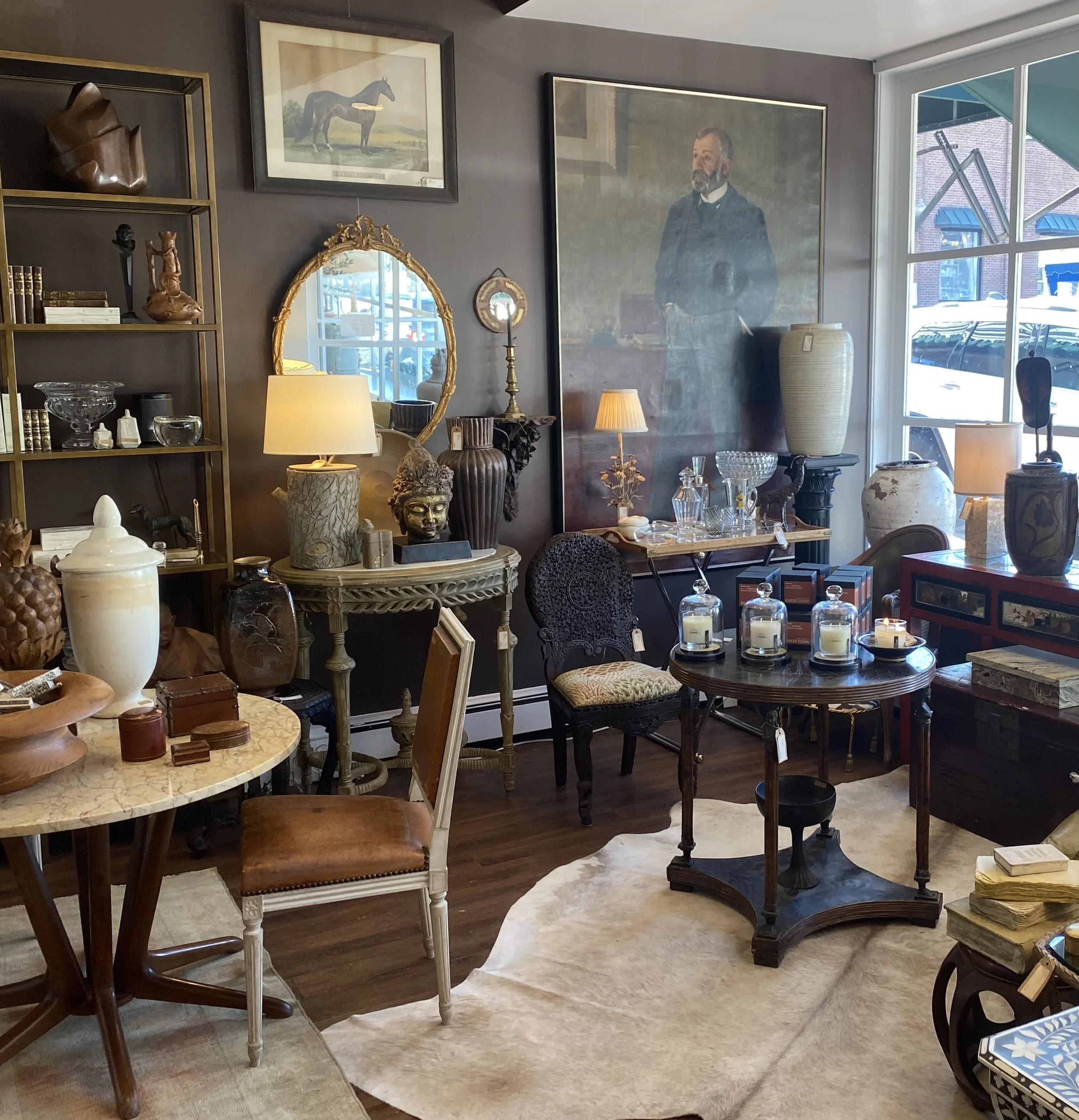 Inside an antique shop with various furniture and decorative items, including a large portrait of a man in a suit, framed artwork of a horse, a gold ornate mirror, and various vases and sculptures displayed on tables and shelves.