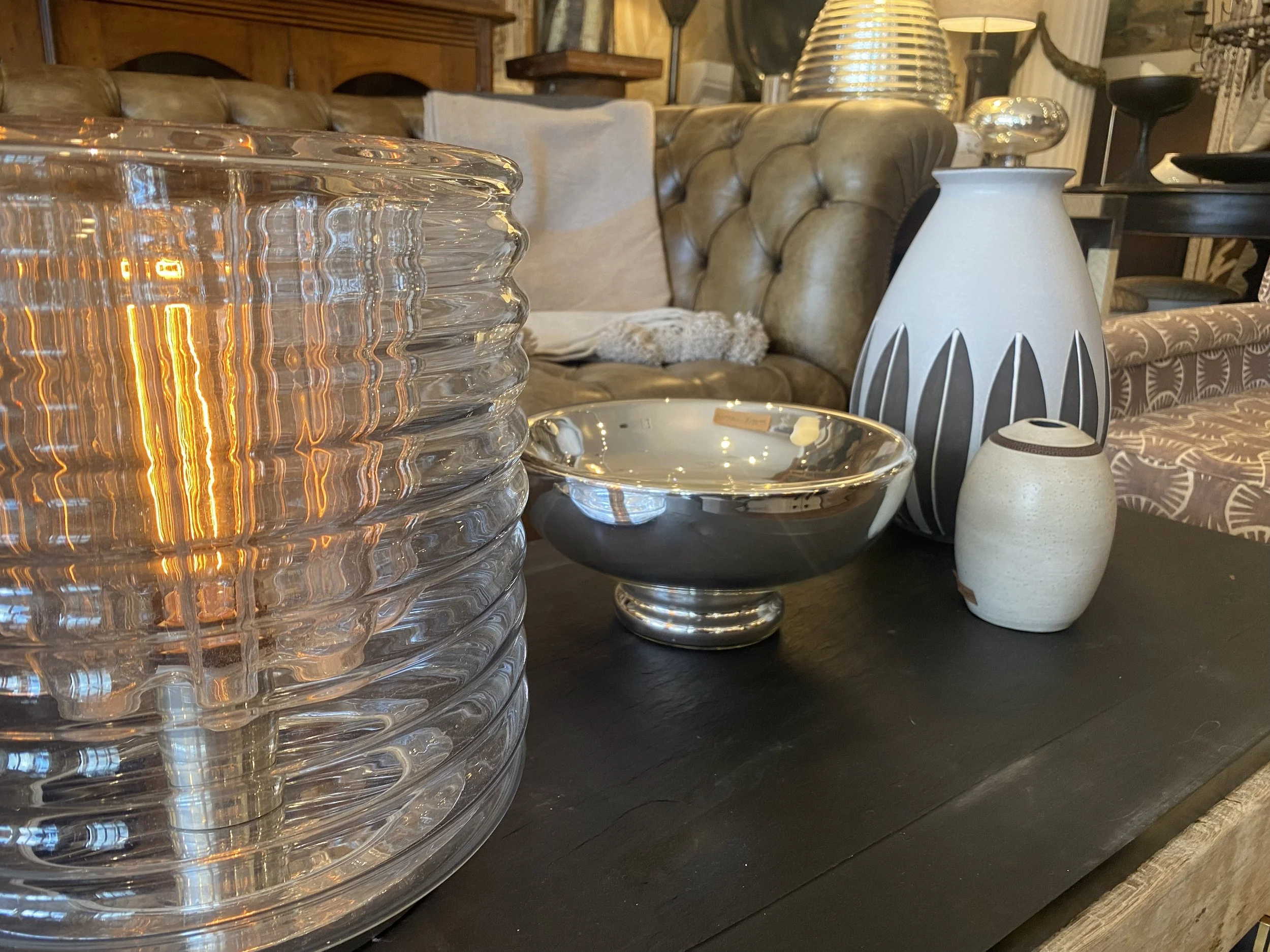 Close-up view of a glass table lamp with a textured, ridged pattern and warm light. In the background, there are various decorative objects, tables, and a couch in a cozy living room setting.