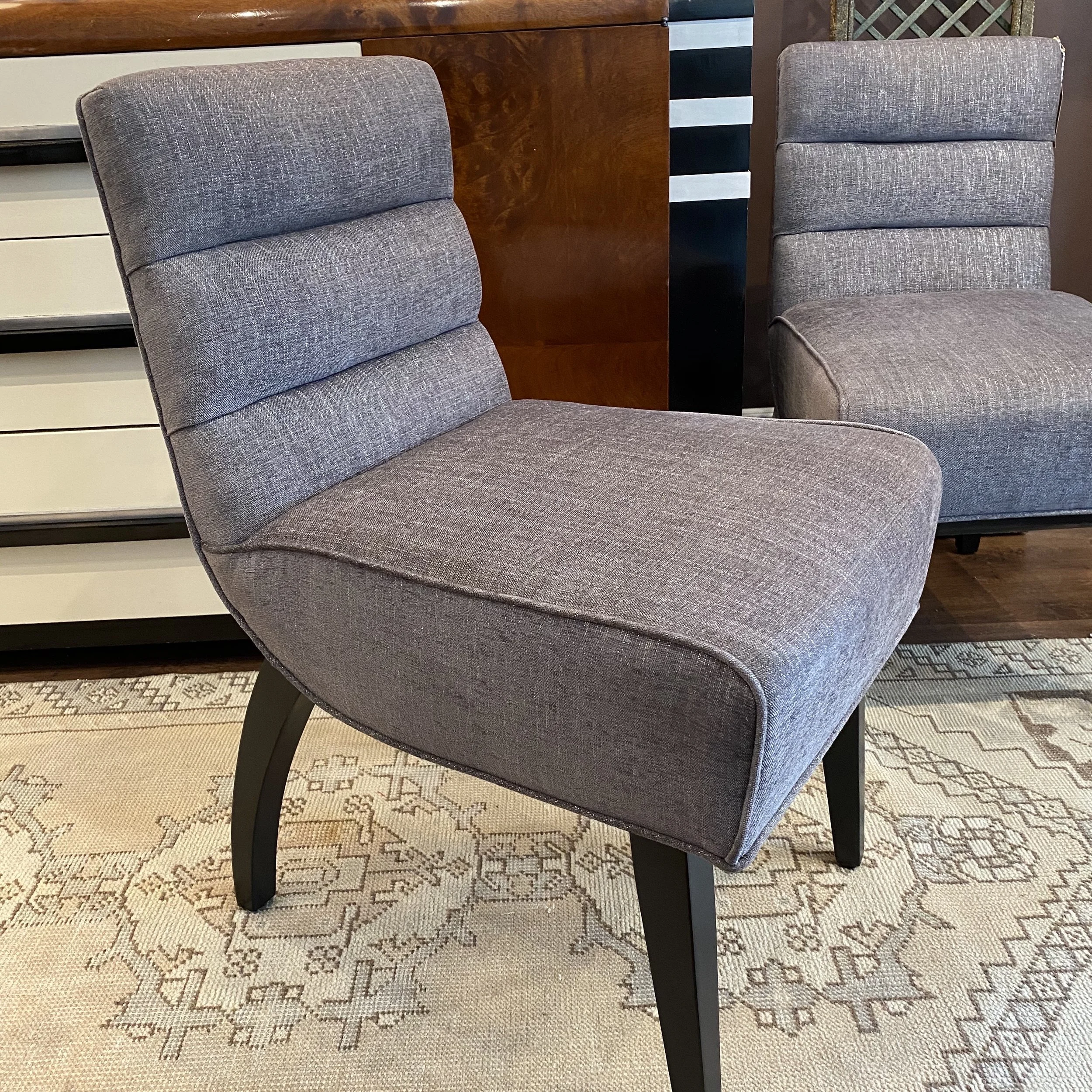 Two gray upholstered chairs with black wooden legs placed on a decorative rug, with a wooden dresser and a black and white striped box in the background.