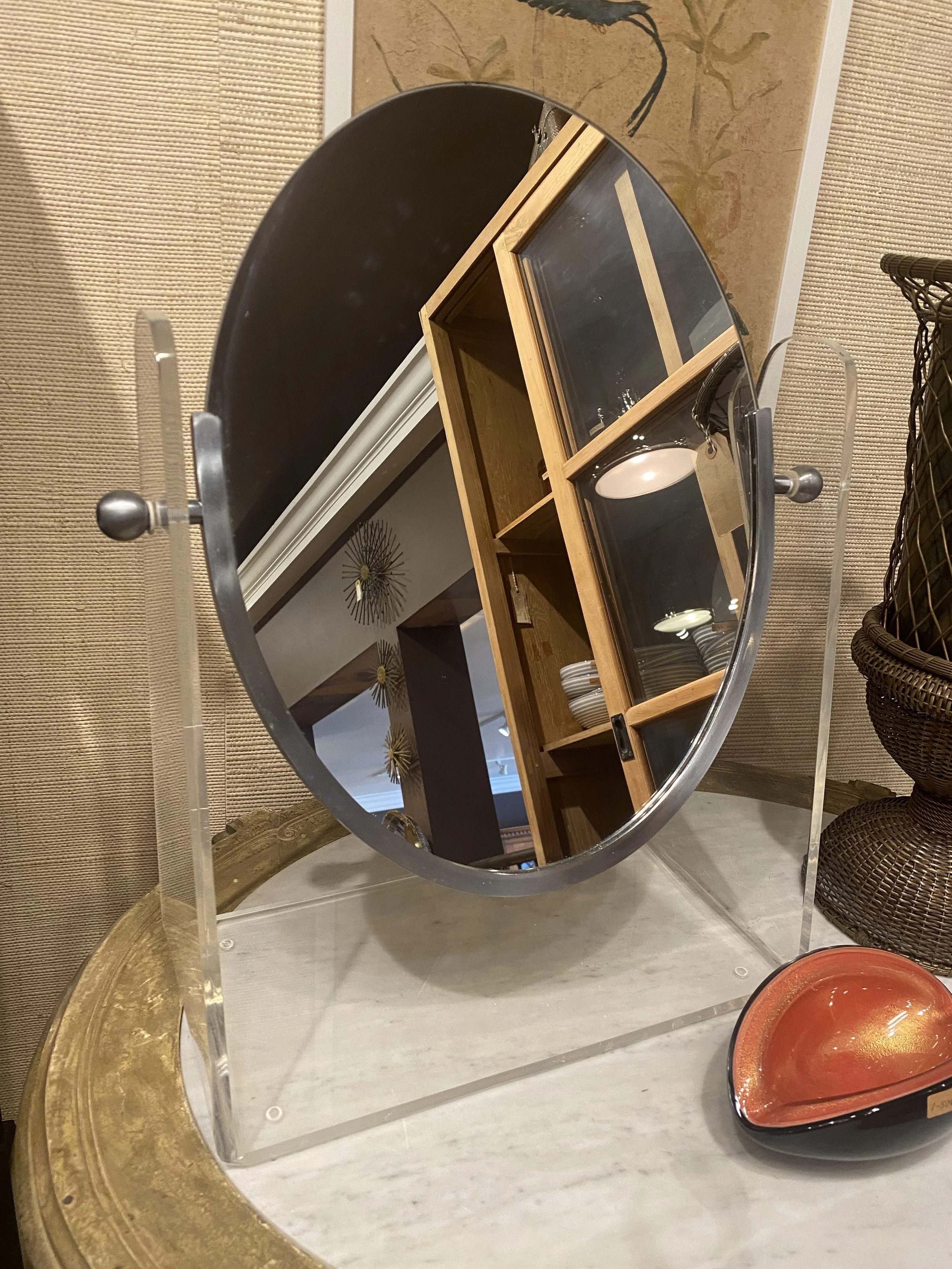 A display mirror with a curved shape showing reflections of a wooden cabinet with glass doors, bowls, and a decorative wall piece, set on a gold-colored tray, next to a basket and other items.