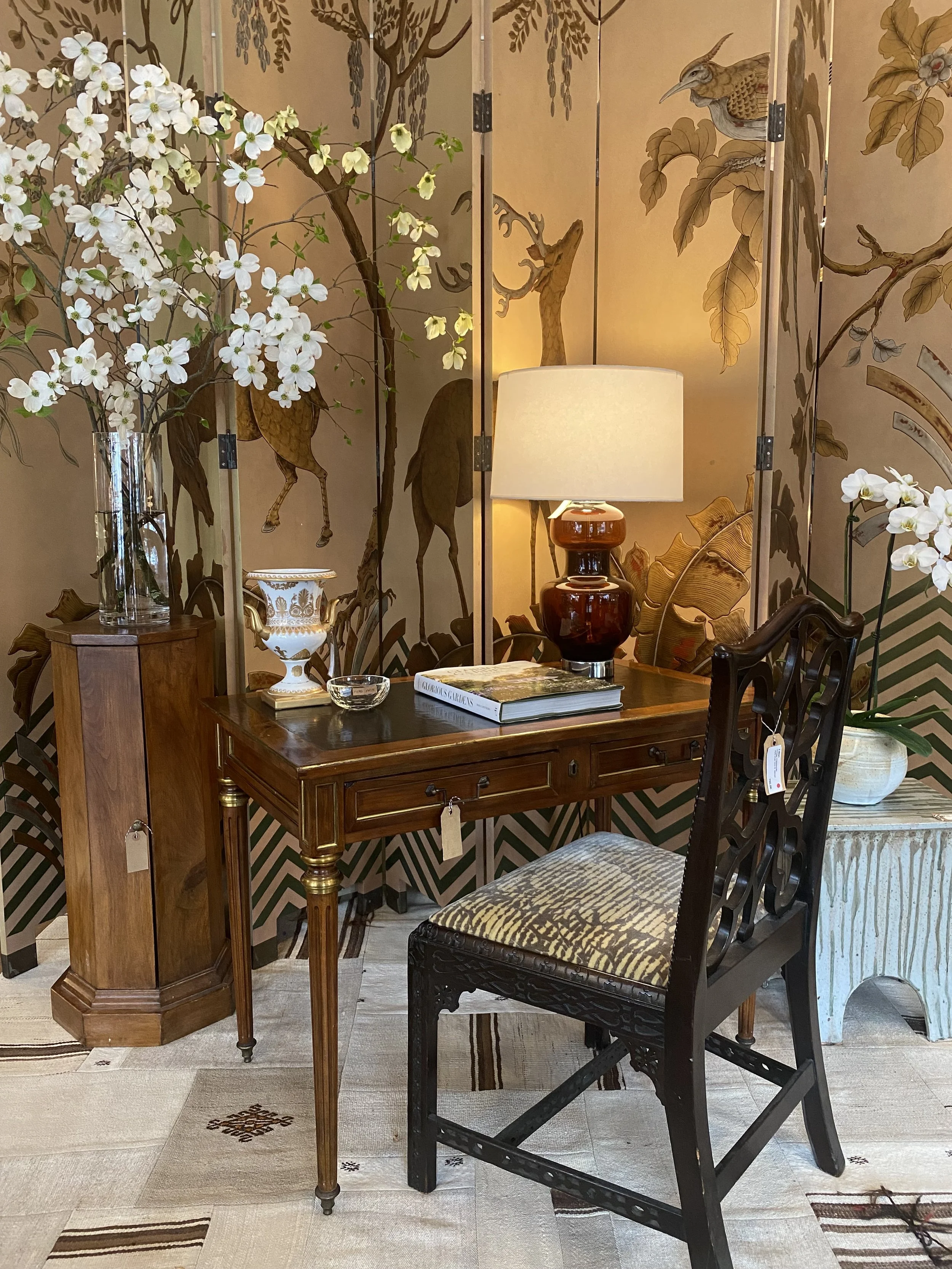 An interior scene with a wooden table, a black chair with animal print fabric, a white lamp on a brown vase, and a decorative screen with a nature scene including birds and trees. There are also flowers in a glass vase and a large potted plant.