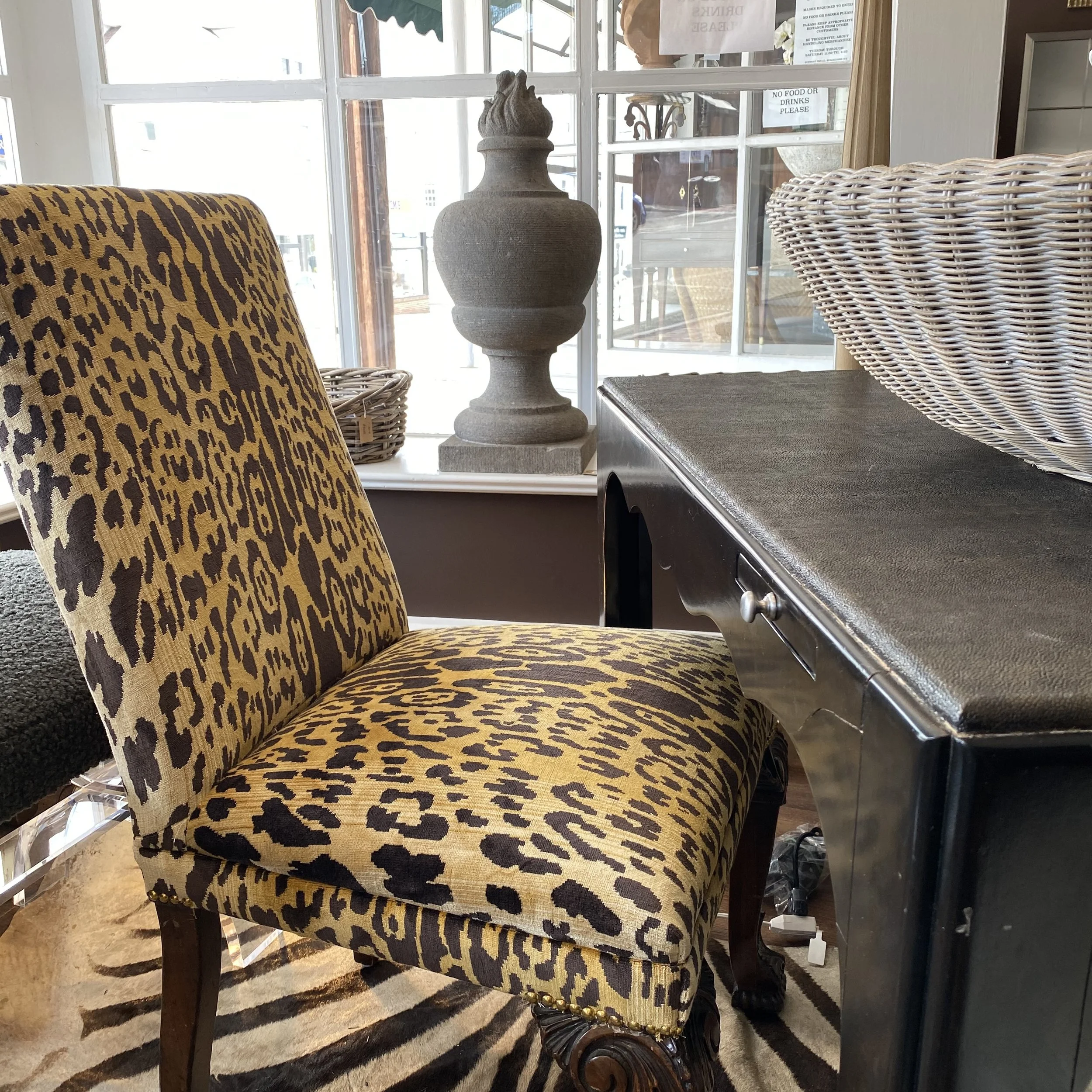 A leopard print upholstered chair with dark wooden legs, positioned in a room with large windows. Next to the chair is a wicker basket and a gray stone decorative object resembling a large urn. The chair's back and seat are visible, with the pattern 