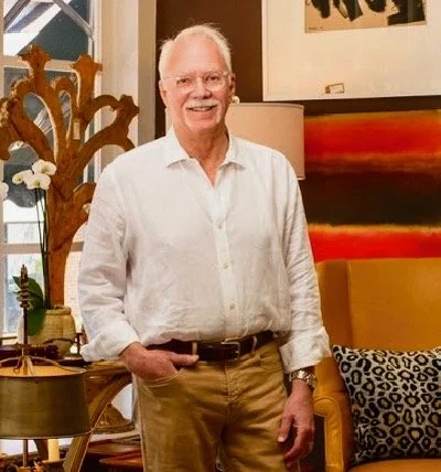 A man with white hair and a mustache stands indoors wearing a white button-down shirt and khaki pants, smiling with one hand in his pocket. He is surrounded by warm-toned furniture, artwork, decorative mirrors, and home décor in a well-appoint