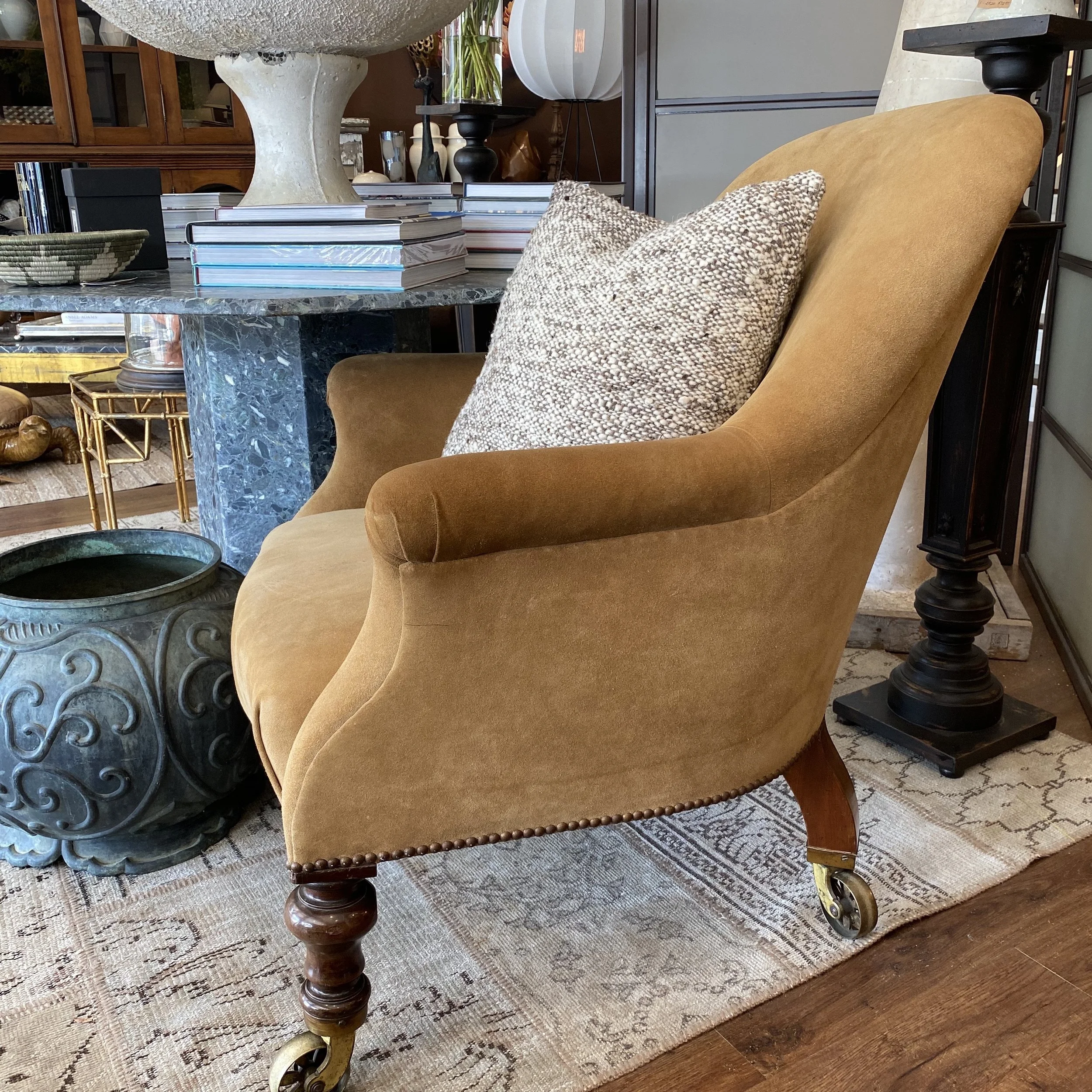 A table with a textured dark surface decorated with a large white stone bowl, stacks of books, and vases with plants. A mustard-yellow upholstered chair with wooden legs and a patterned pillow is partially visible in the foreground. The room has hard