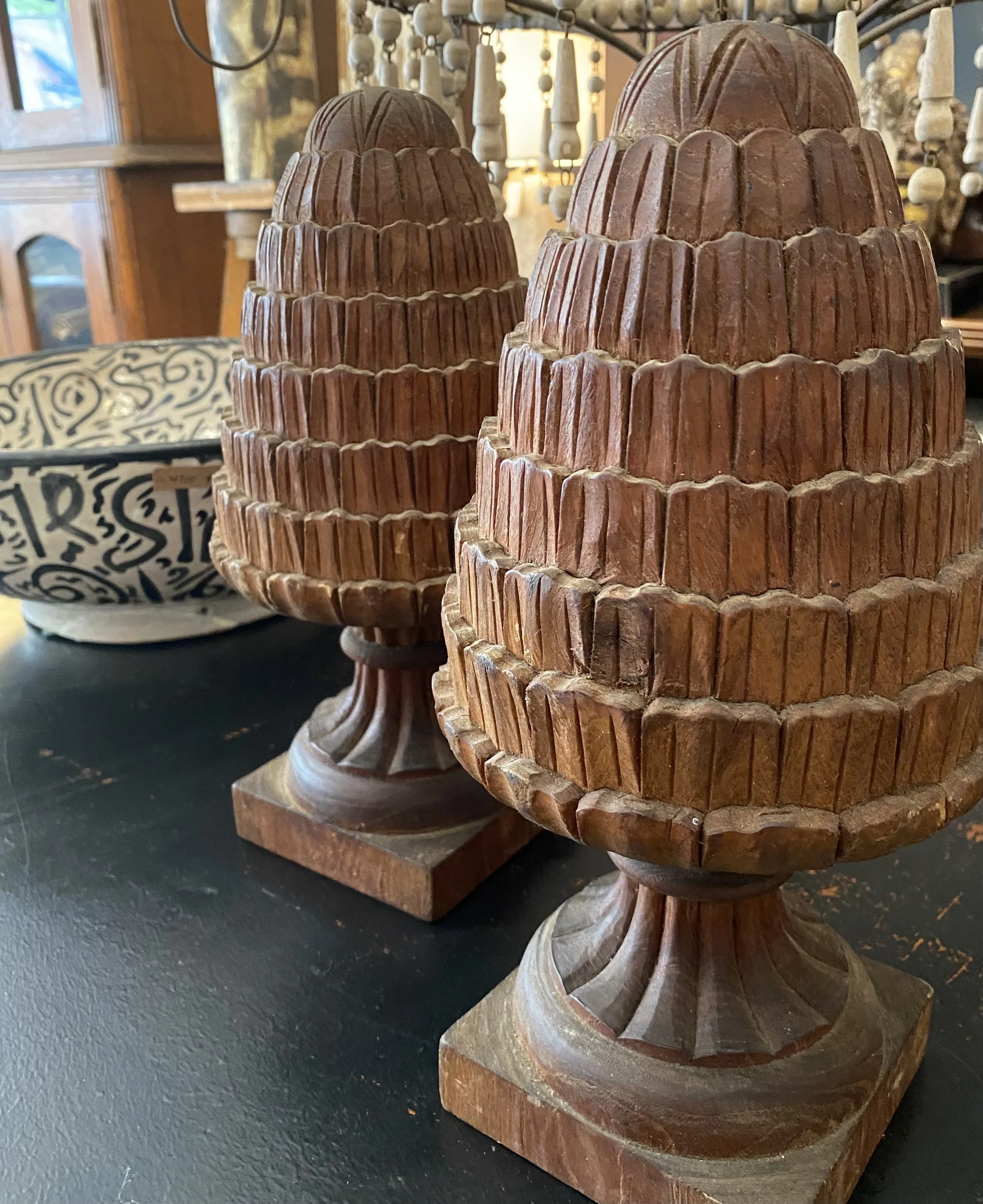 Close-up of a wooden carved sculpture with multiple tiered, rounded, decorative sections, placed on a carved wooden base. Other similar sculptures and decorative items are visible in the background.