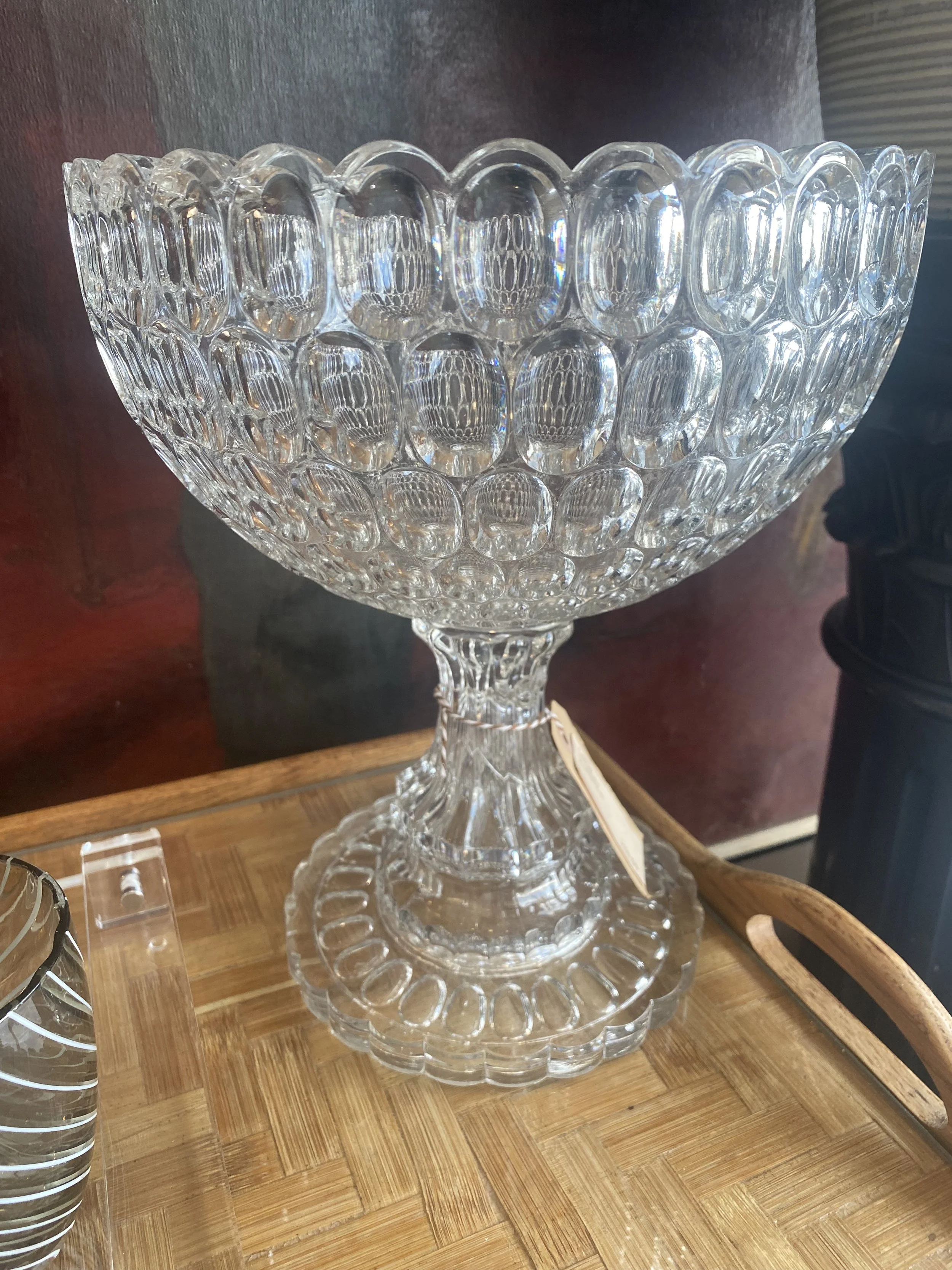 A clear glass bowl with a textured surface, sitting on a wooden surface. The bowl is large and decorative, with an intricate pattern of oval shapes. Blurred objects are nearby, and the background has a dark wall with a red tint.