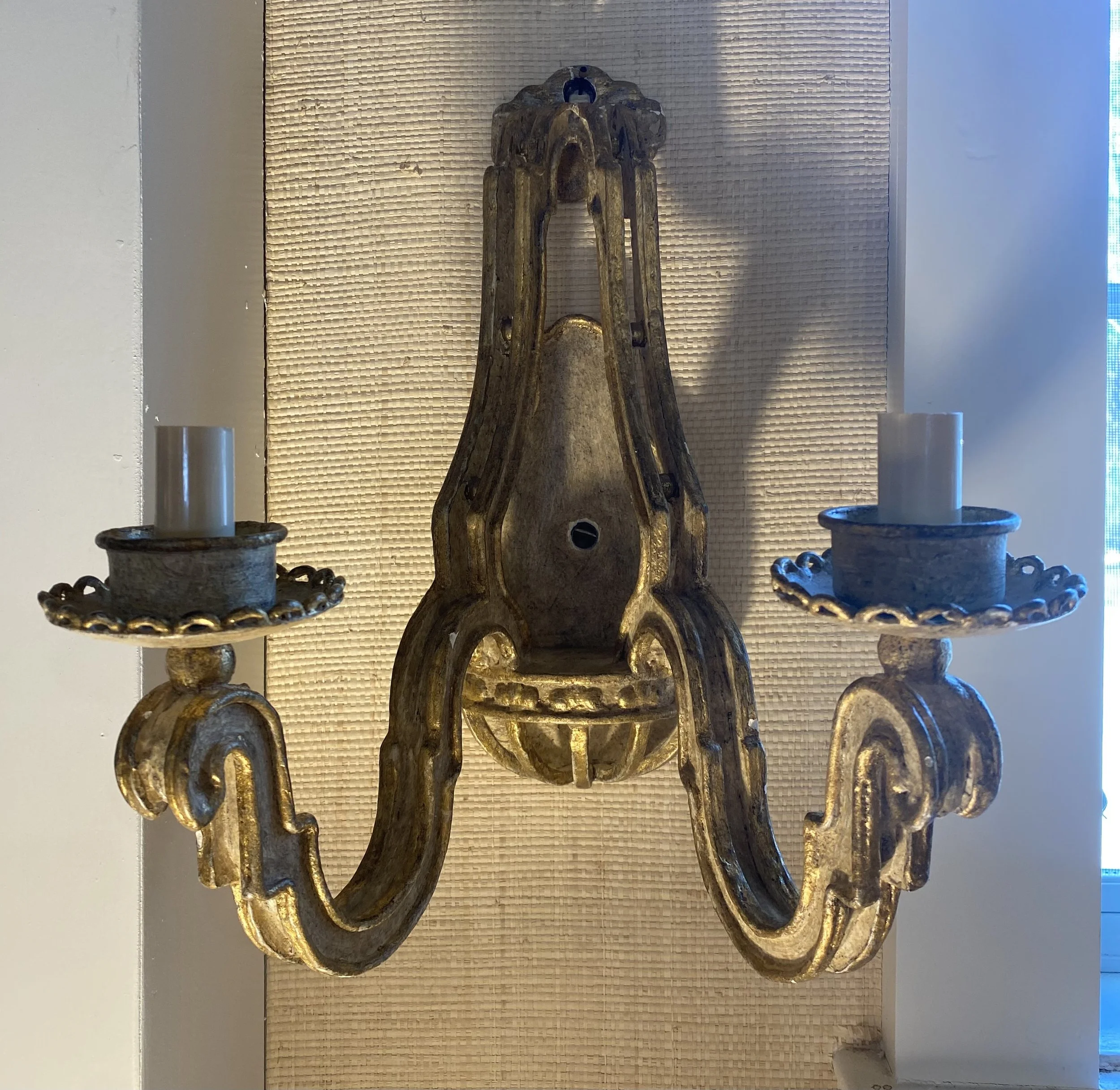 Decorative wooden sculpture flanked by metal candle holders with candles, set against a textured beige wall with indirect lighting.