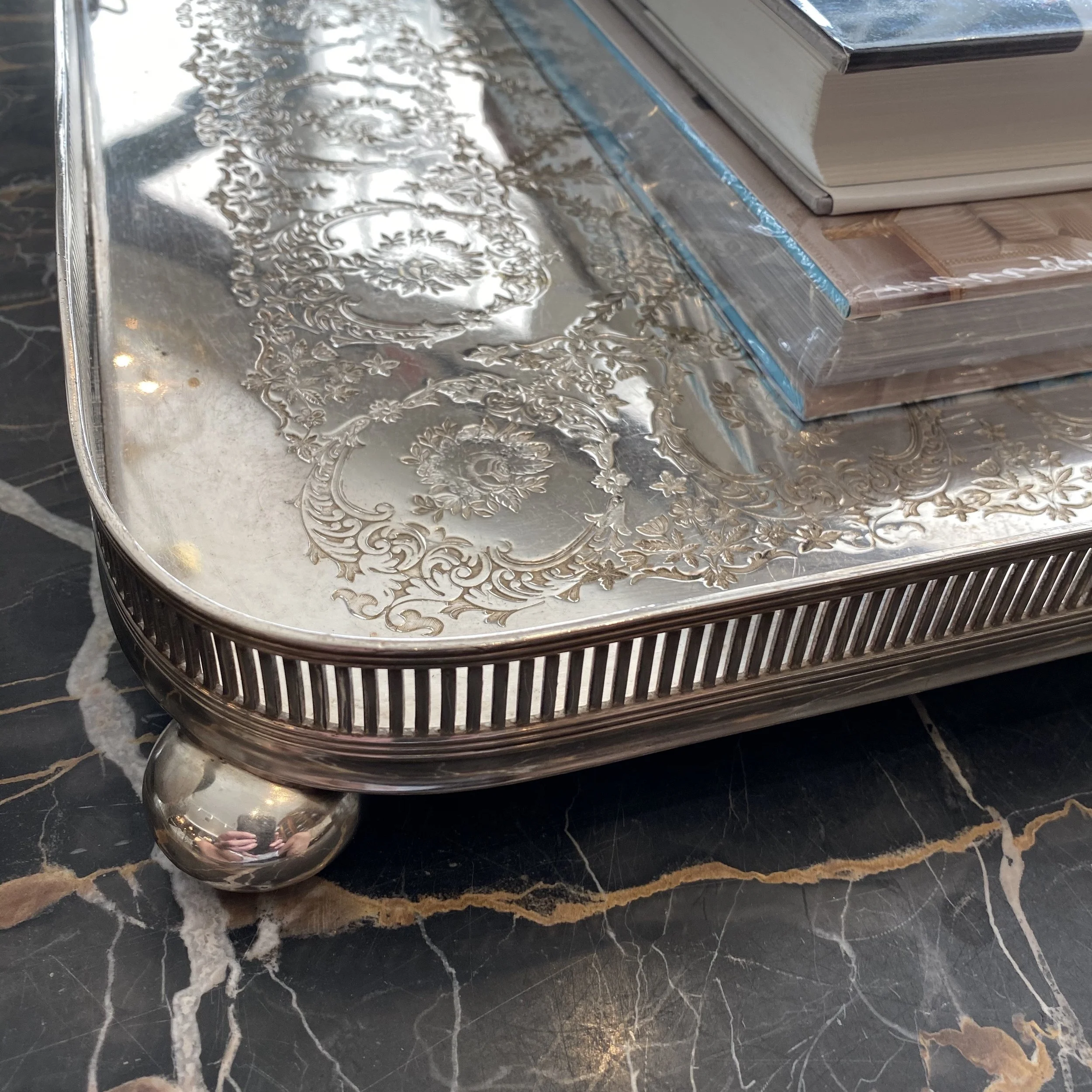 A silver ornate decorative metal tray placed on a black and brown marble surface, with stacked hardcover books in the background.