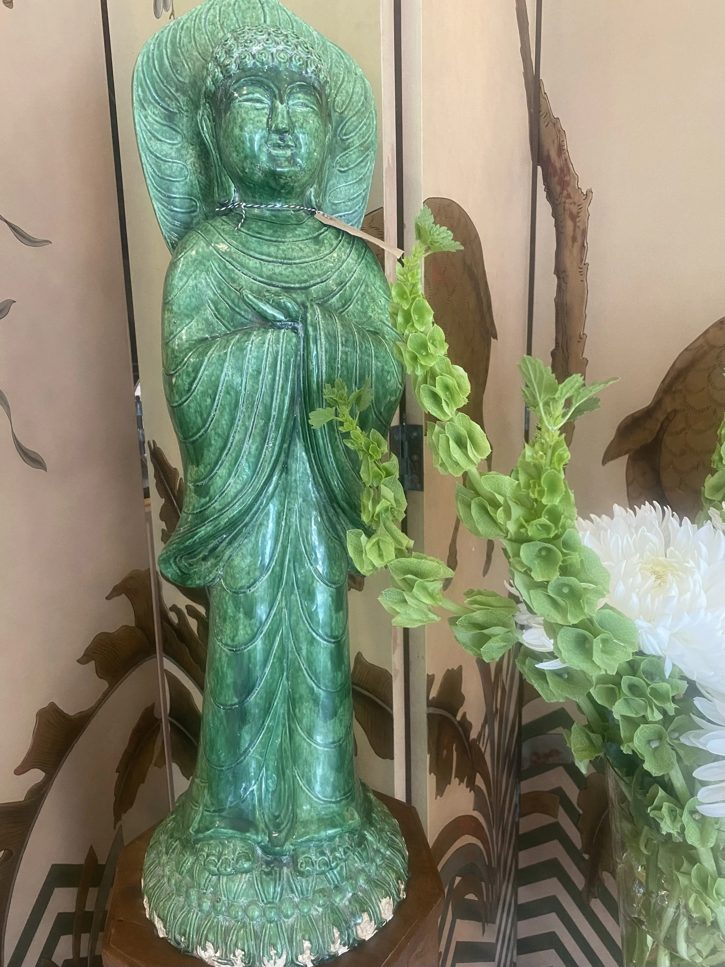 A large green Buddha statue with closed eyes, seated with hands in lap, decorated with carvings and patterns. The statue is surrounded by artificial green leaves and white flowers. A decorative backdrop with dark wooden leaves and a checkered floor c