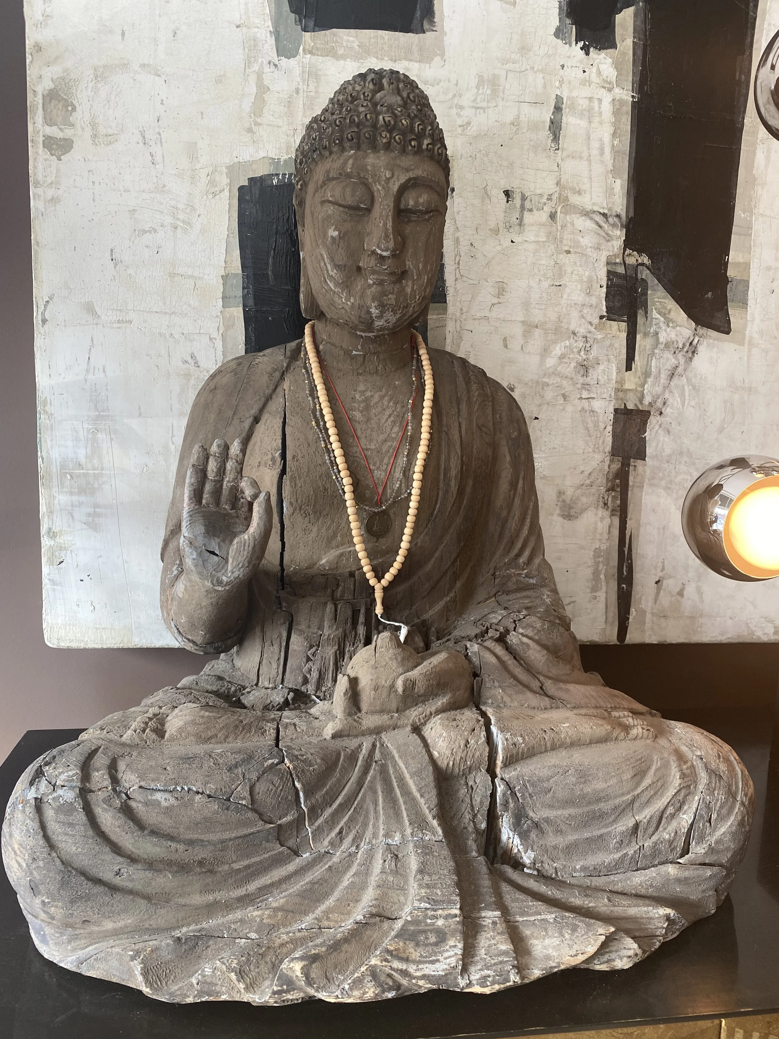 A weathered wooden sculpture of a seated Buddha with a serene expression, wearing necklaces, and making a hand gesture, set against a textured wall.