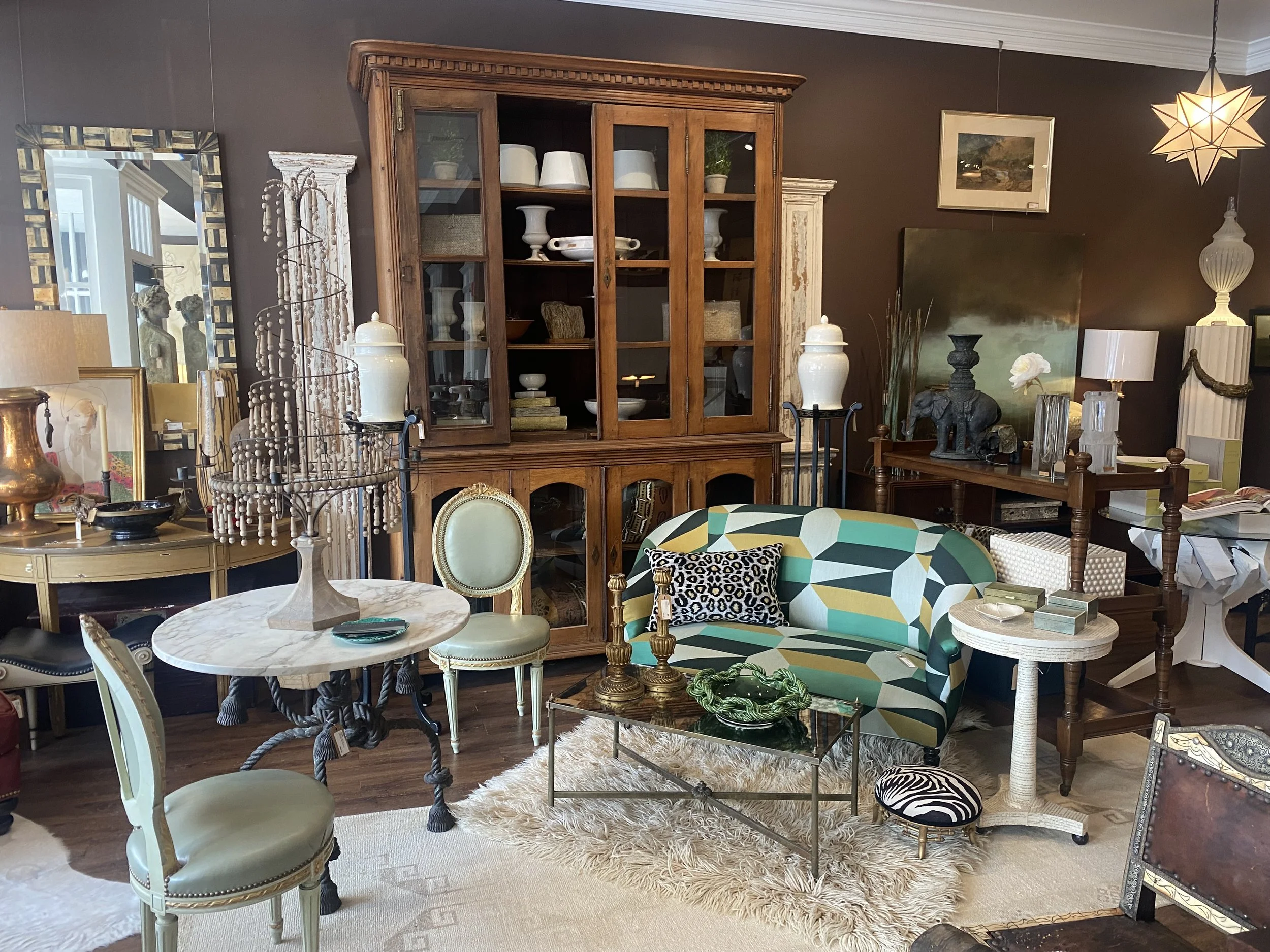 Interior of a furniture store showcasing a variety of furniture pieces including wooden cabinets, lamps, decorative objects, a sofa with a geometric pattern, a round marble table, and framed artwork on the walls.