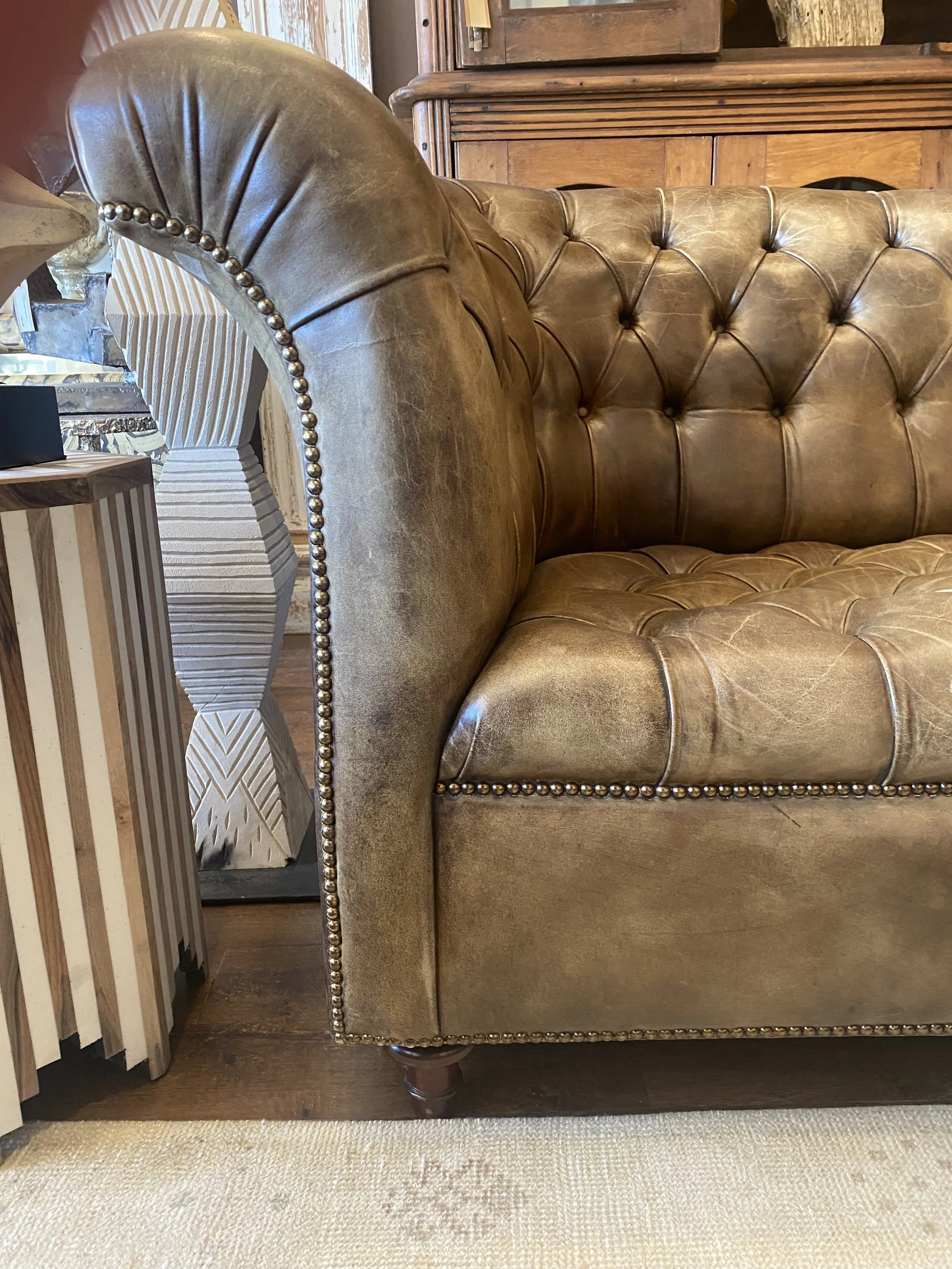 A close-up photo of a vintage tufted leather sofa with brass nailhead trim on a wooden floor, with a beige carpet in the foreground and a striped box and wooden furniture in the background.