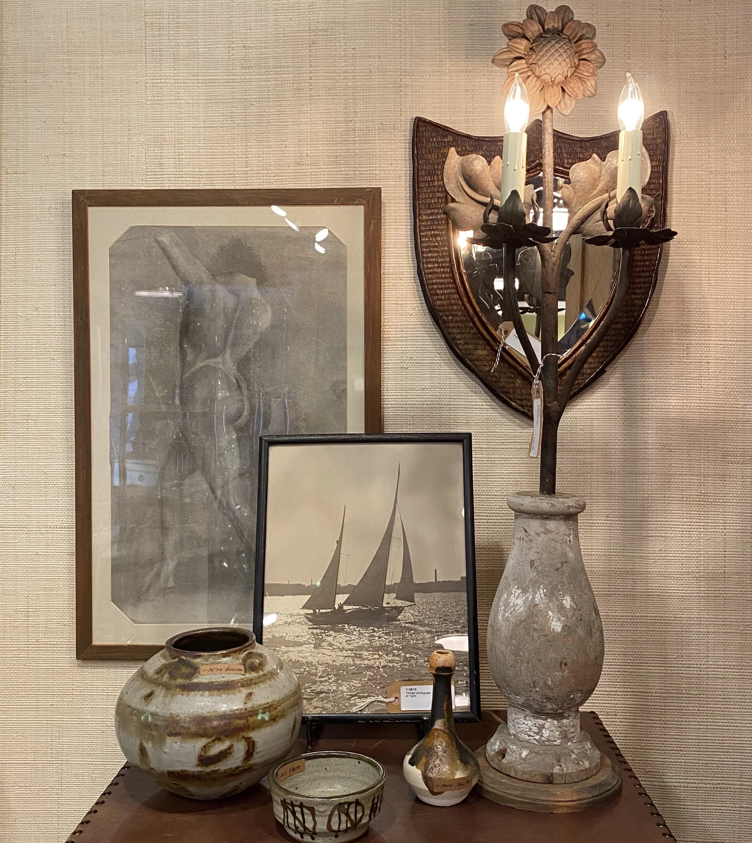 Display of various ceramic and pottery objects, including vases and bowls, arranged on a table, with framed artwork and a decorative wooden wall piece with lights in the background.