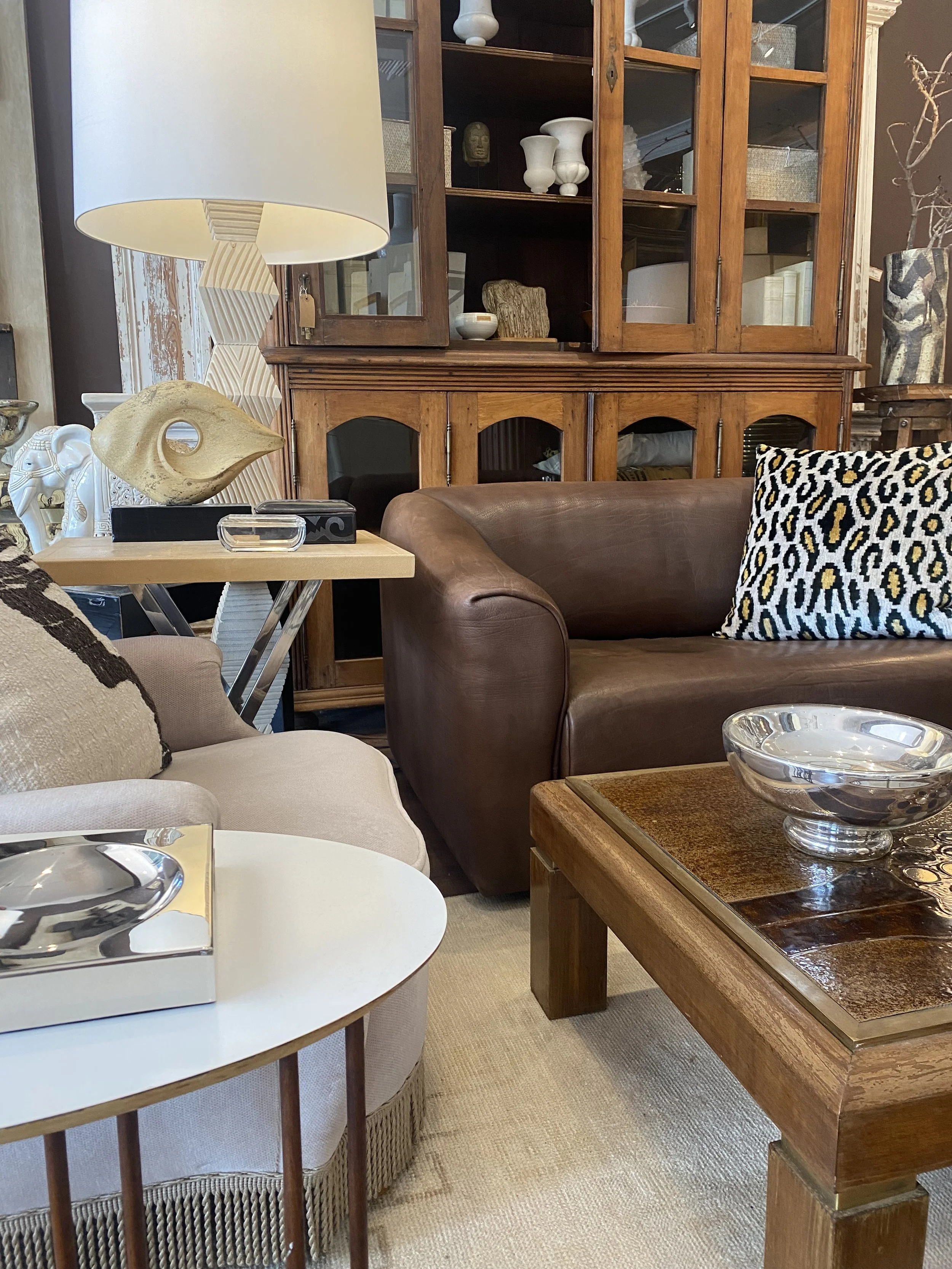 Living room with a wooden cabinet, a brown leather sofa with a decorative pillow, a round white coffee table with a decorative tray, a lamp with a geometric patterned base and white shade, and various decorative items including sculptures and vases.