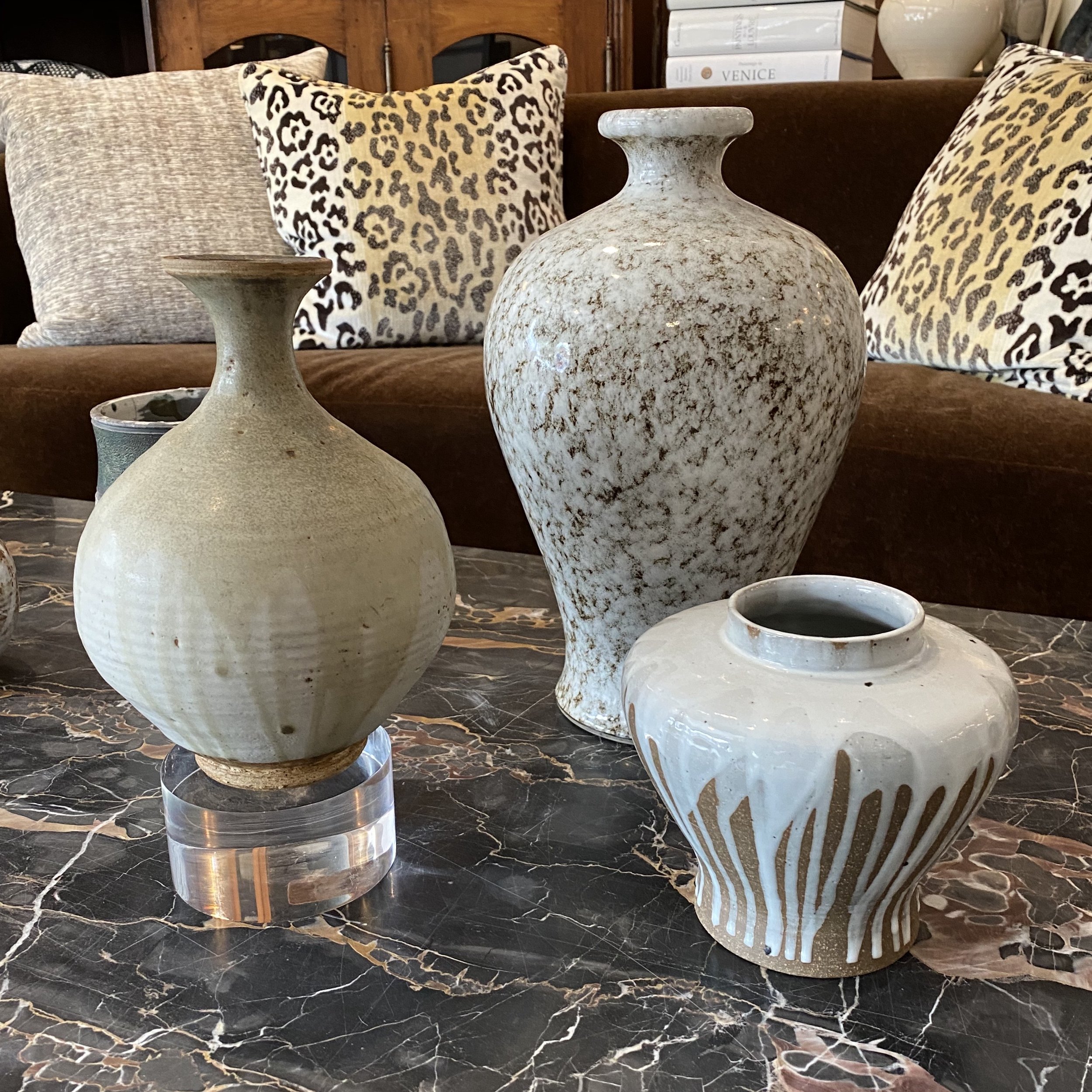 Several marble vases of different shapes and sizes placed on a black and brown marbled surface, with pillows in animal print and a wooden cabinet in the background.